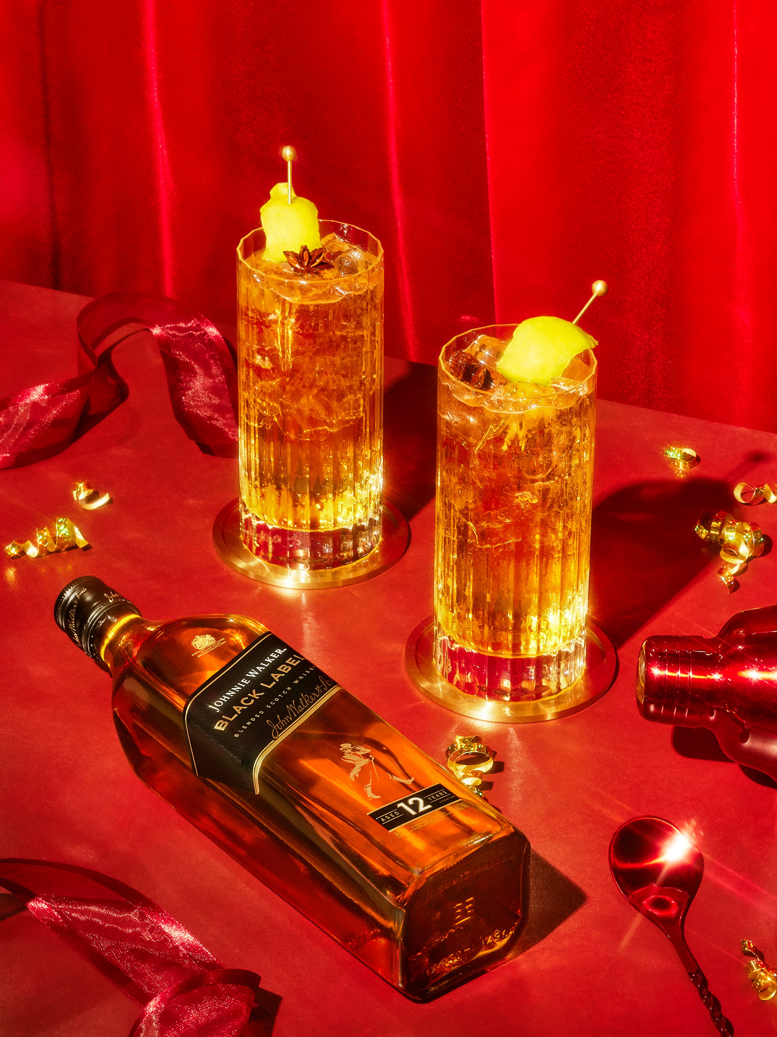 Johnnie Walker Black Label lying beside two highball cocktails on a red set with ribbon and confetti