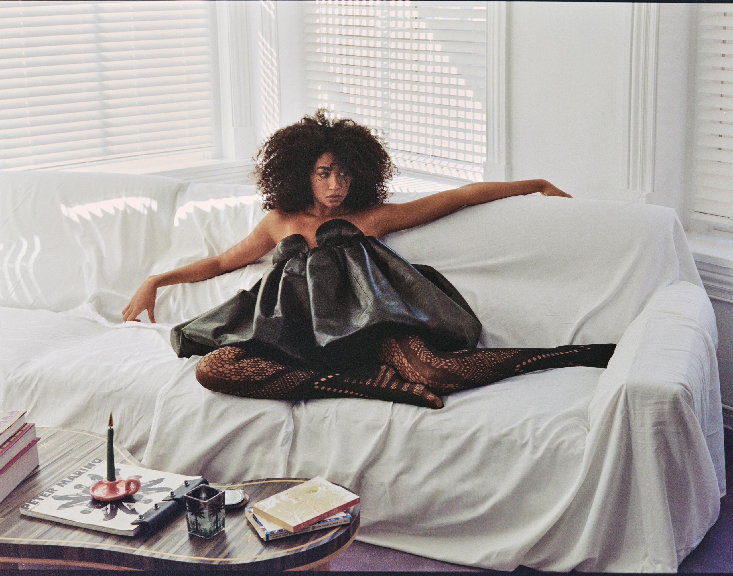 Aoki Lee Simmons stretched across a sheet-draped white sofa in a sculptural black leather dress and patterned tights, window light