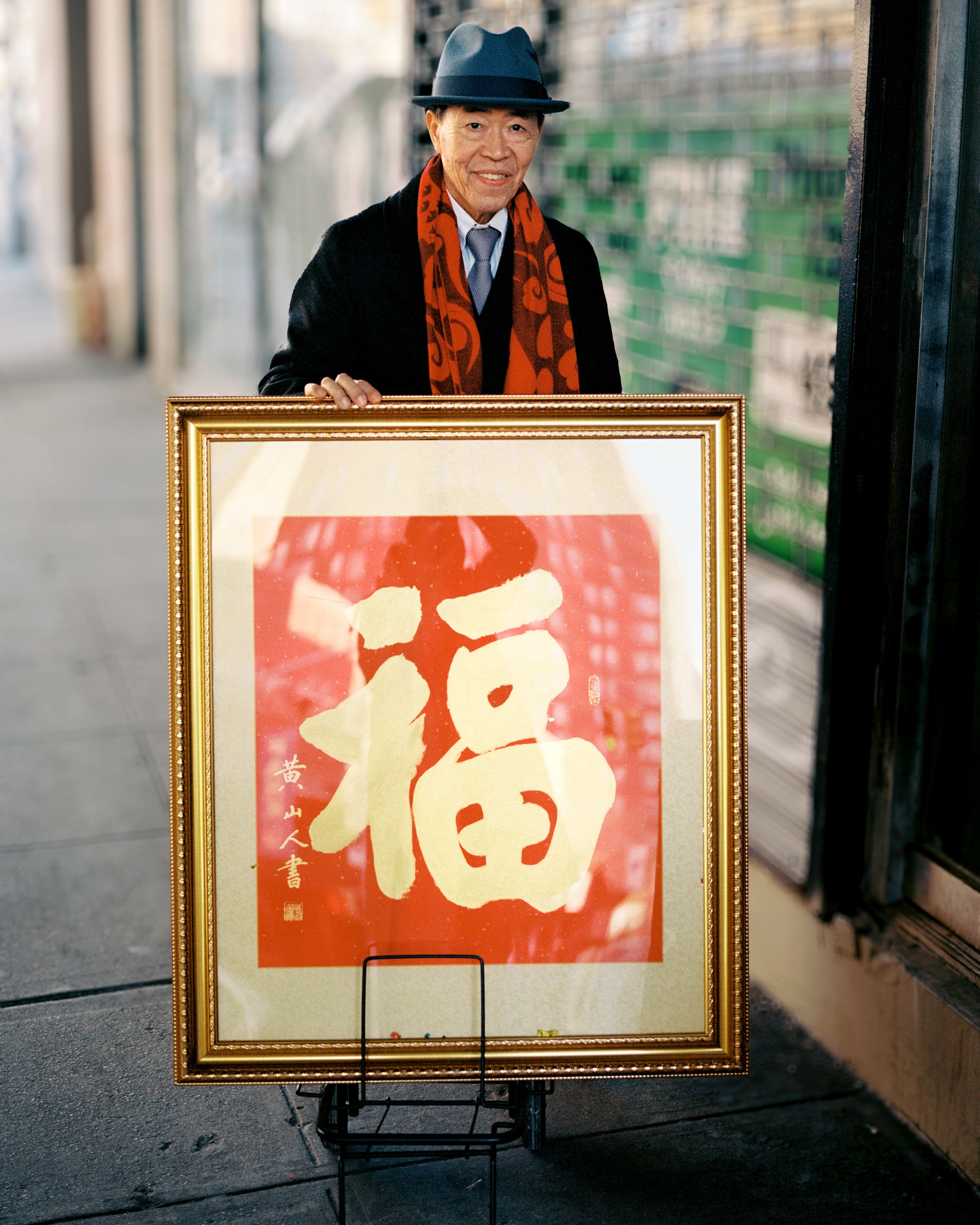 Man in a red scarf beside a framed red fu character, the symbol for good fortune