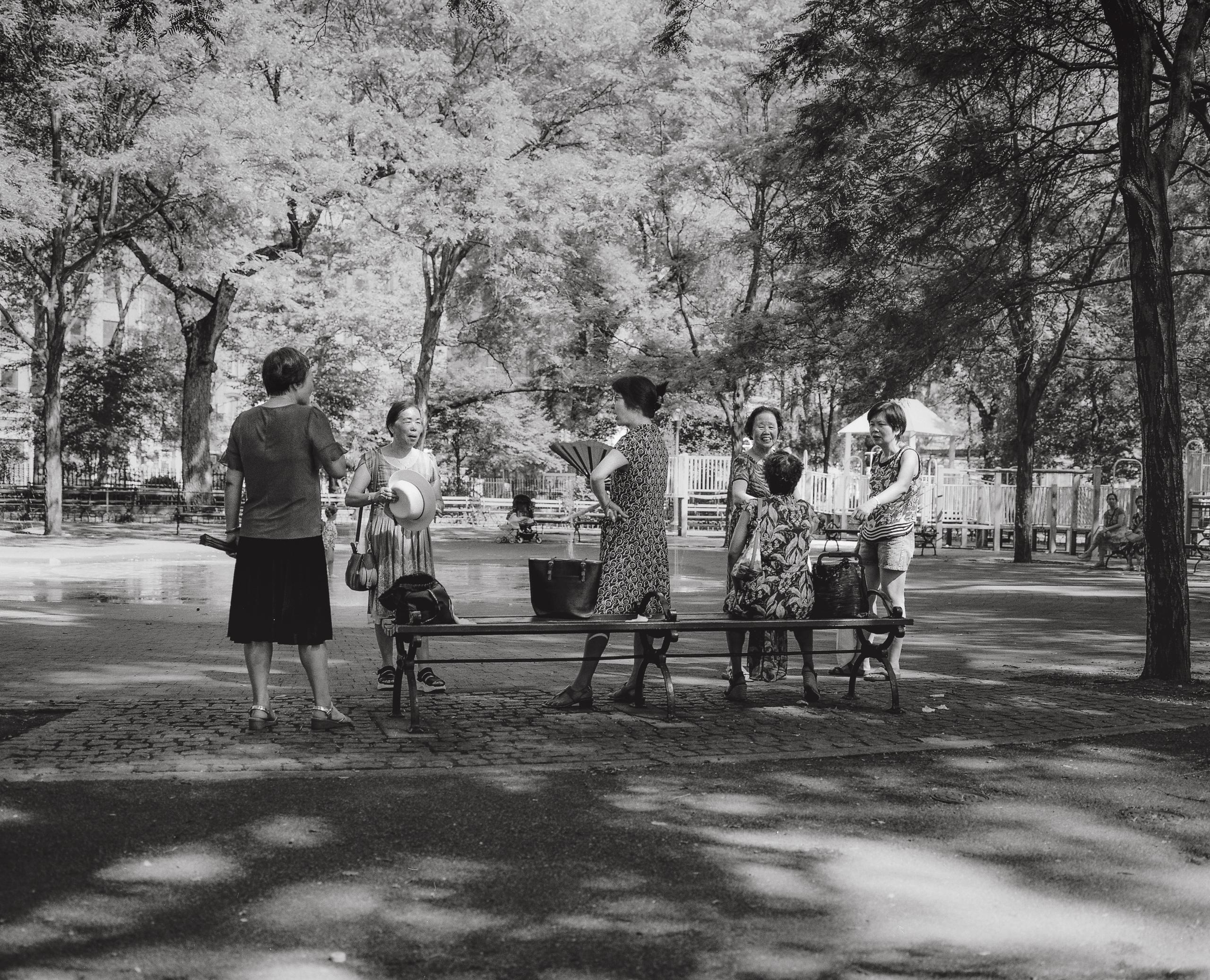 Neighbors gathered around a park bench under dappled shade in Chinatown, black-and-white film