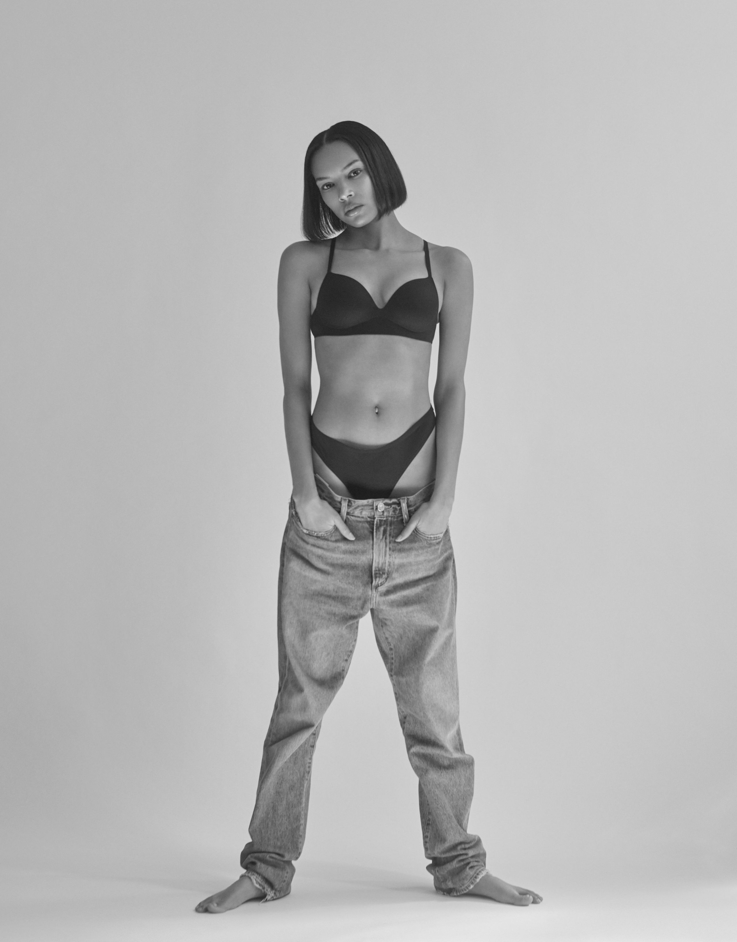 Symon standing barefoot in a black bra and briefs, oversized jeans slung low, hands in pockets