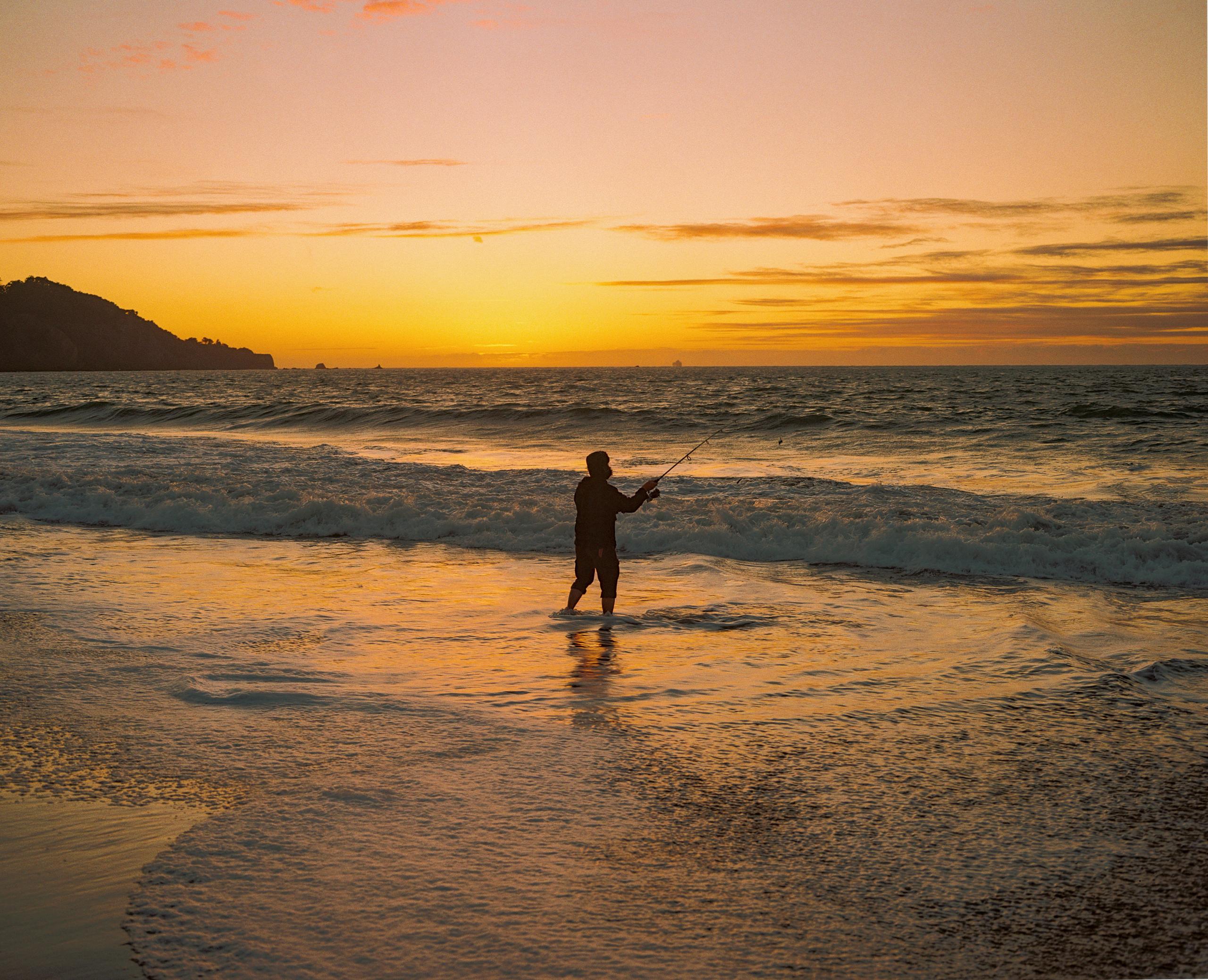 A lone figure standing in the shallows at sunset, the headland dark against a golden sky