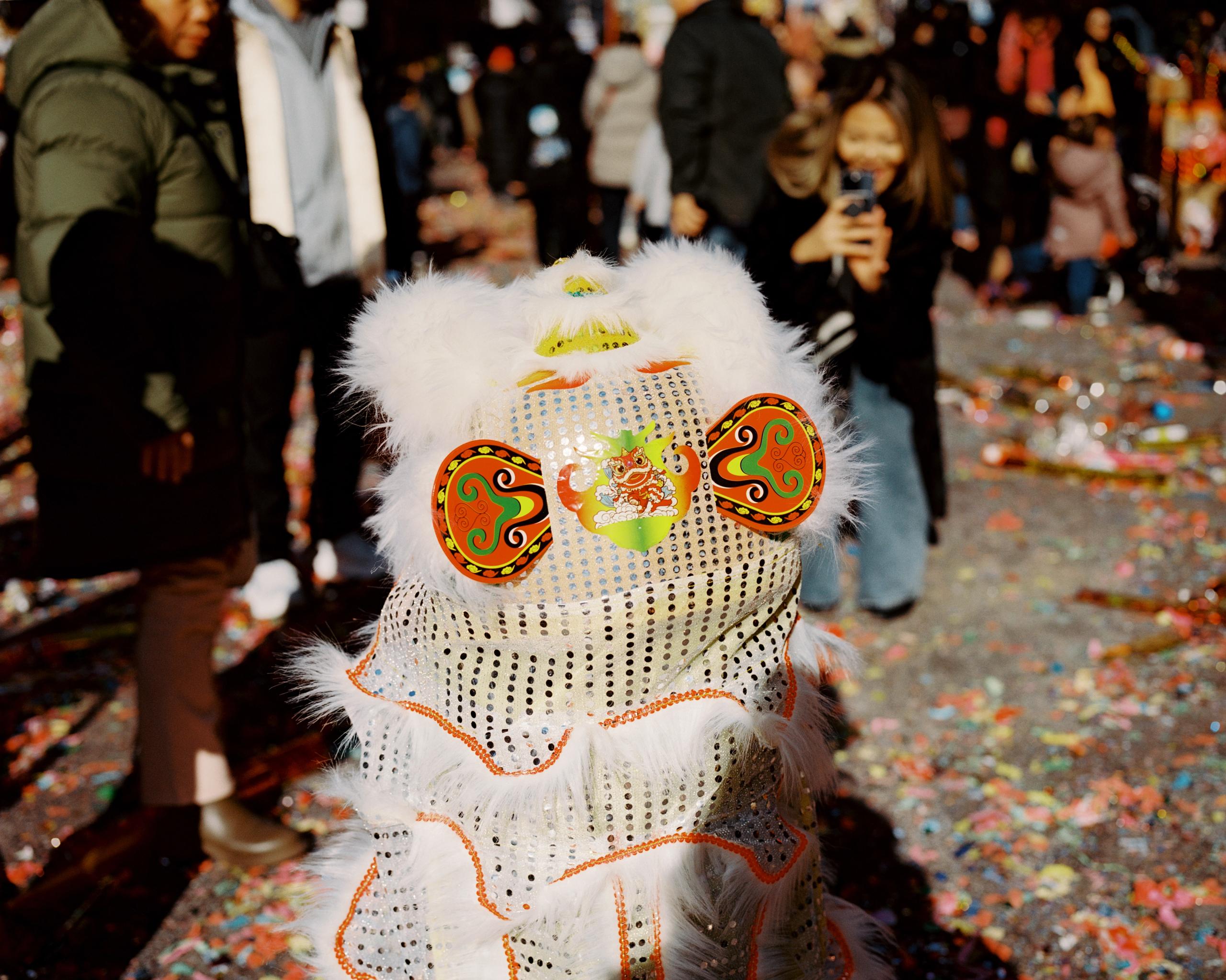 White sequined lion dance head glowing in the sun, confetti on the ground behind