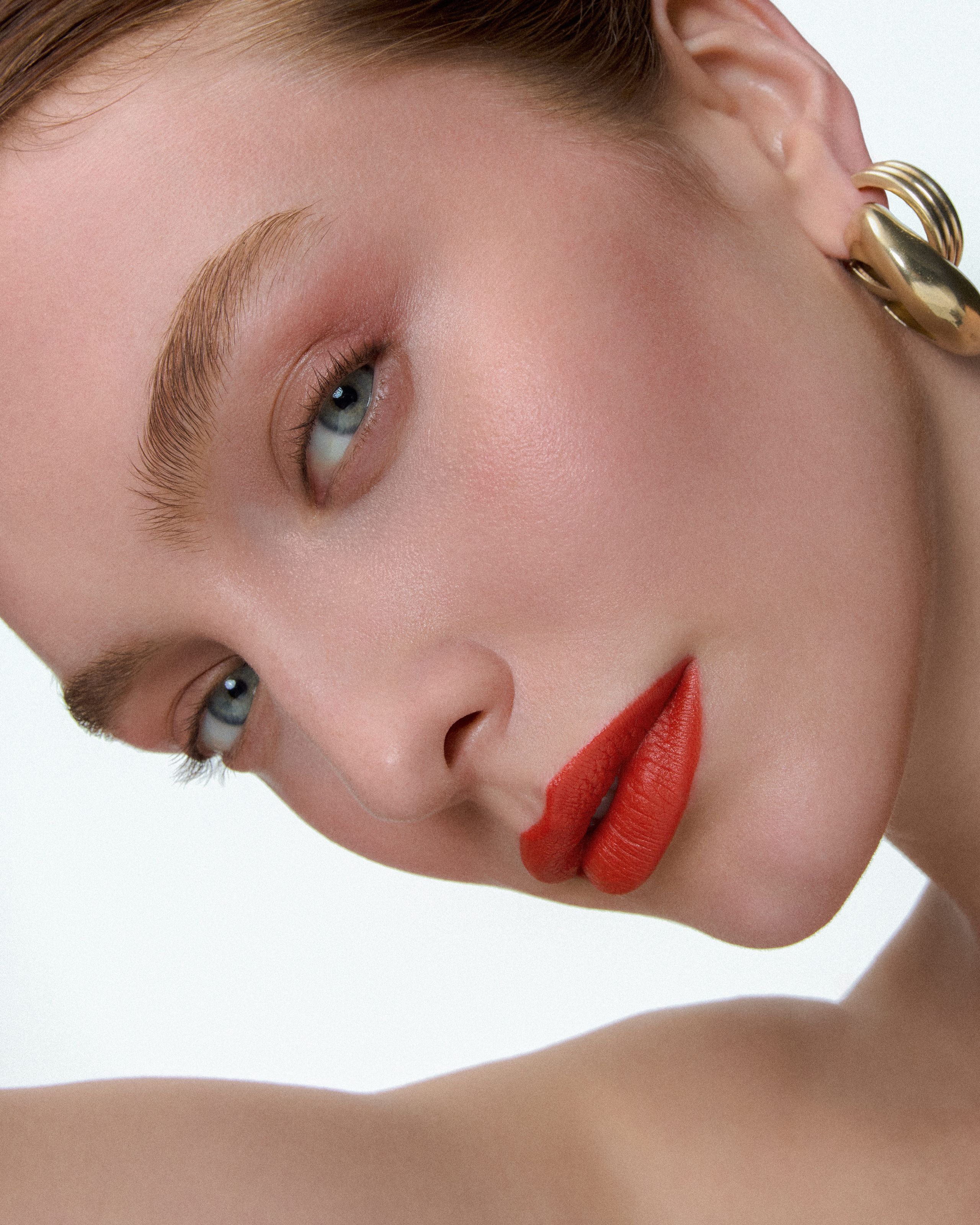 Model resting her head sideways, coral lips and a gold AGMES earring in soft white light