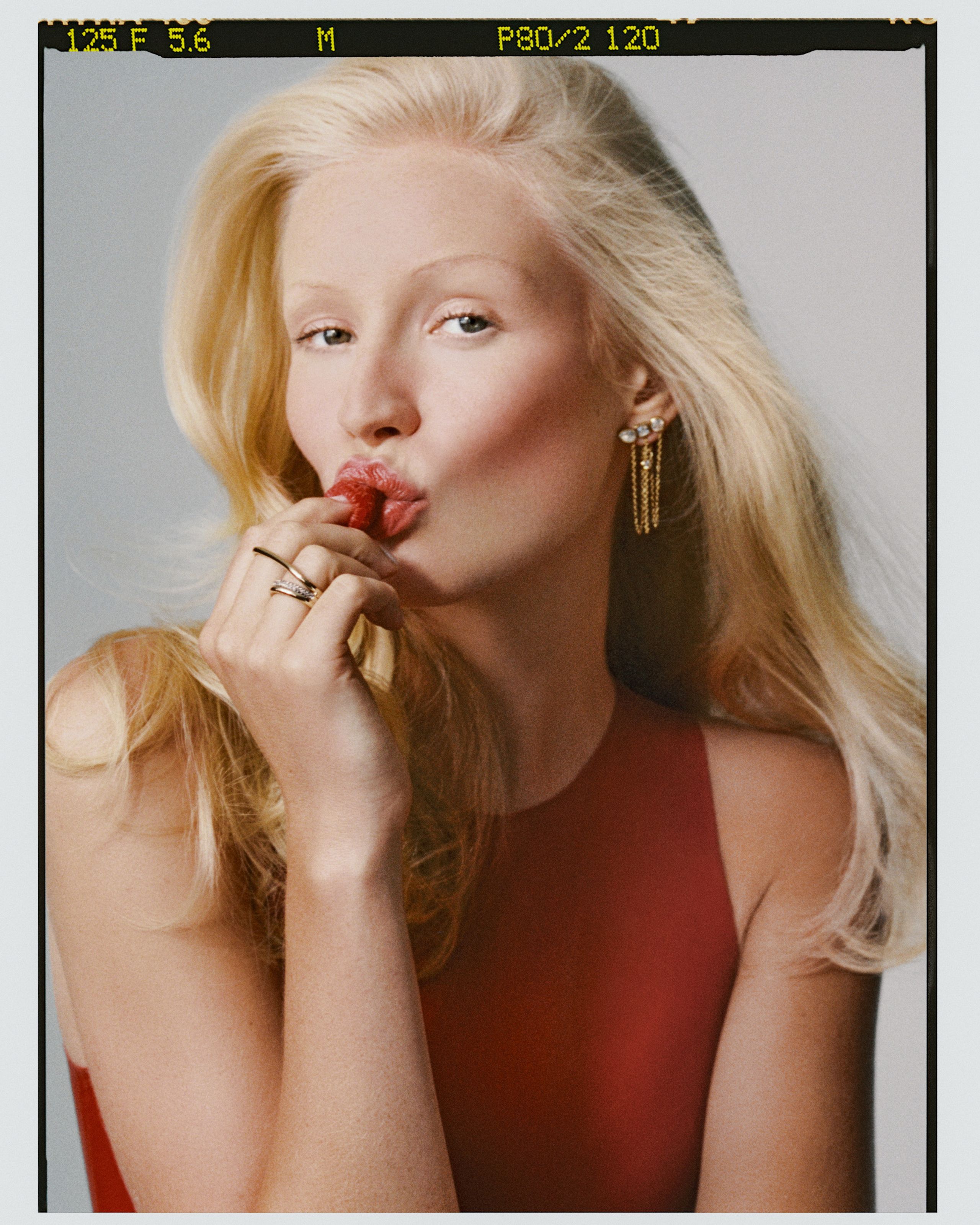Film frame of a blonde model in a rust-red top, fingers to pursed red lips, gold rings and earrings