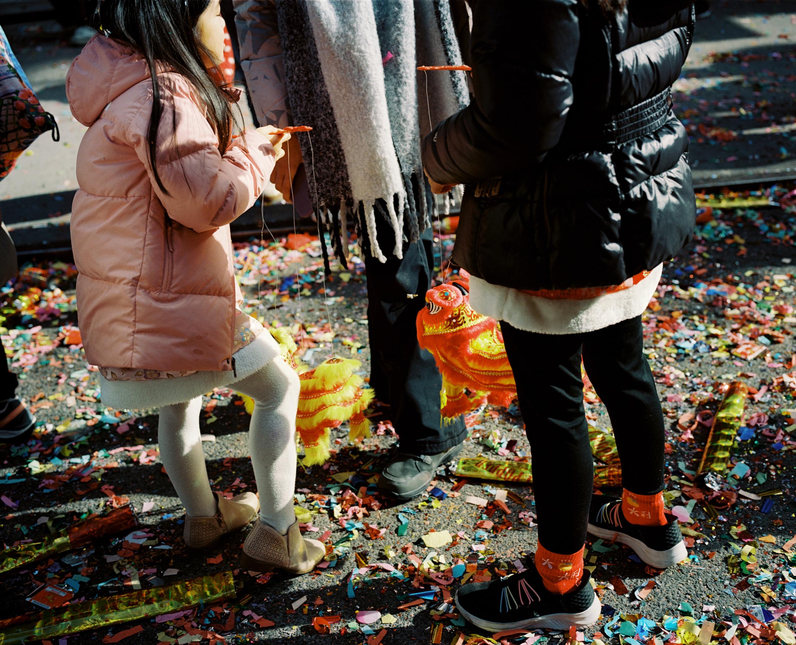 Child in a pink coat standing on a stool above firecracker paper strewn across the street