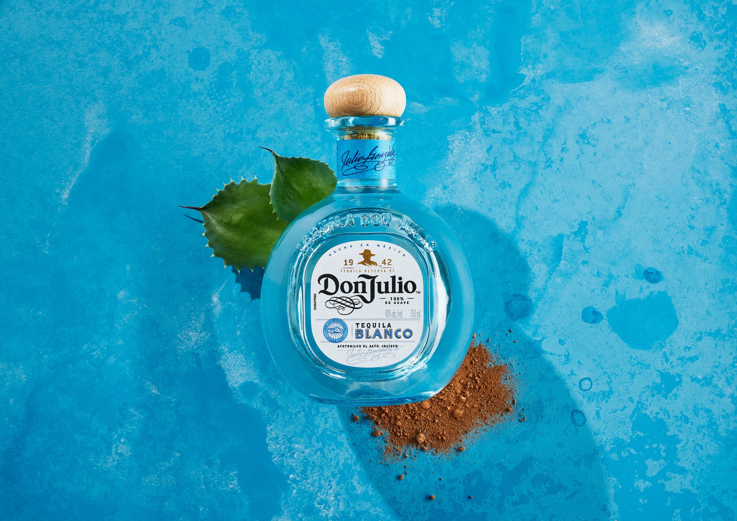 Don Julio Blanco on a watery blue surface with an agave leaf and a scattering of red spice