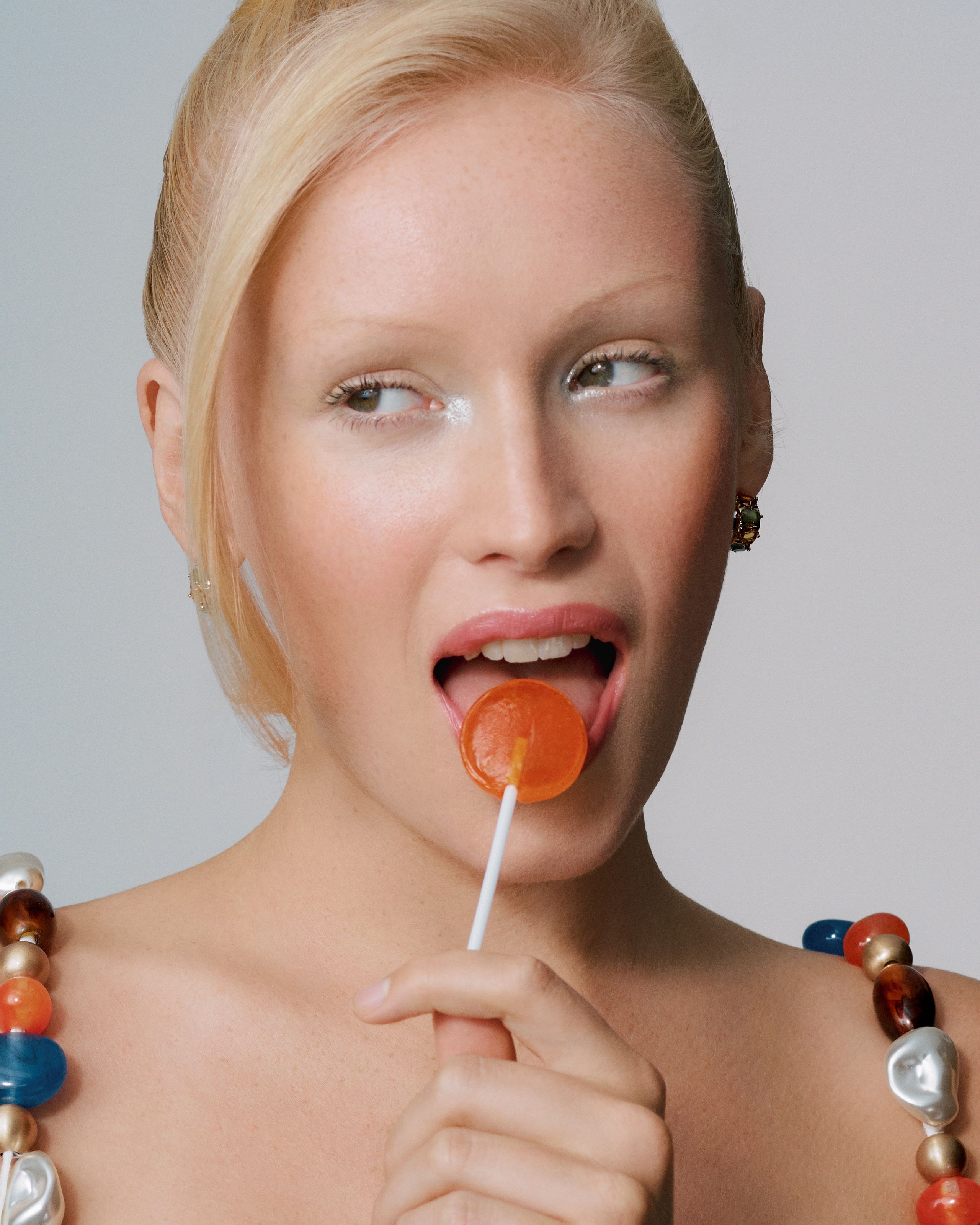 Blonde model licking an orange lollipop with a sideways glance, chunky multicolor bead necklace