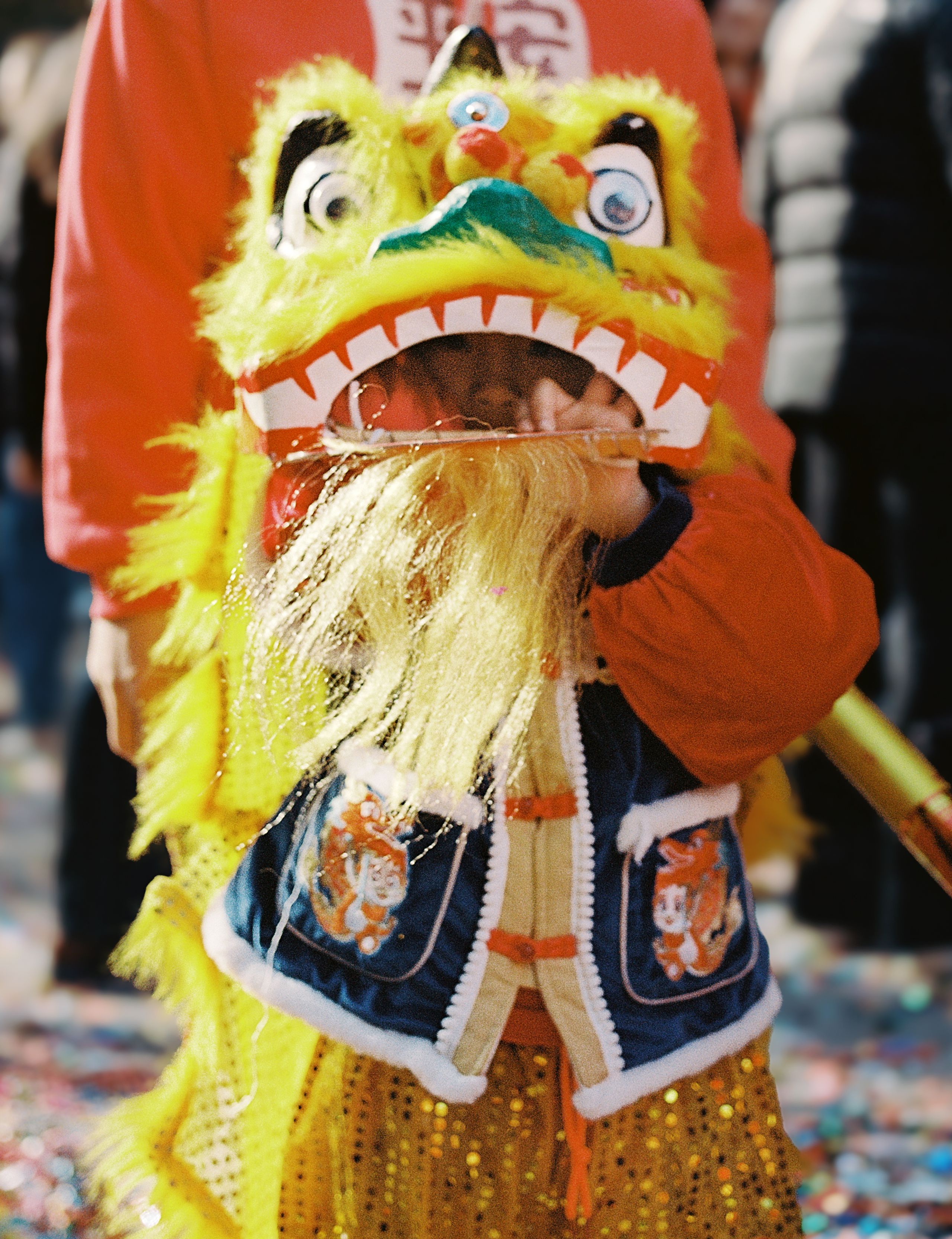 Child wearing a yellow and red lion dance head, confetti scattered at their feet