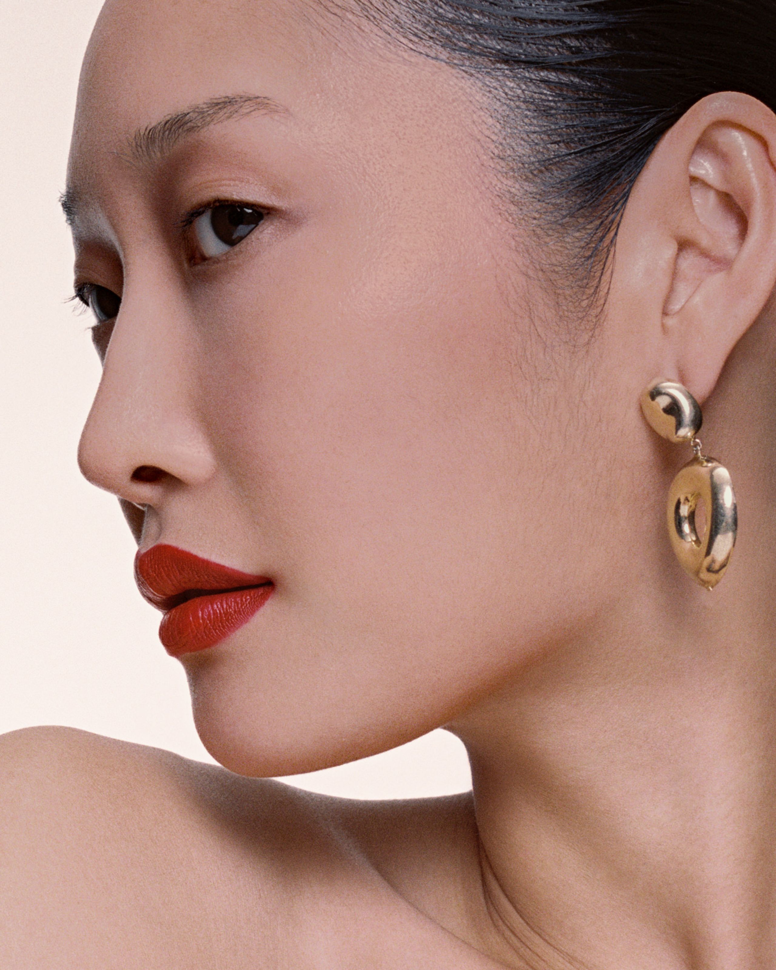 Profile of a model with sleek dark hair, red-orange lips and a gold AGMES disc earring