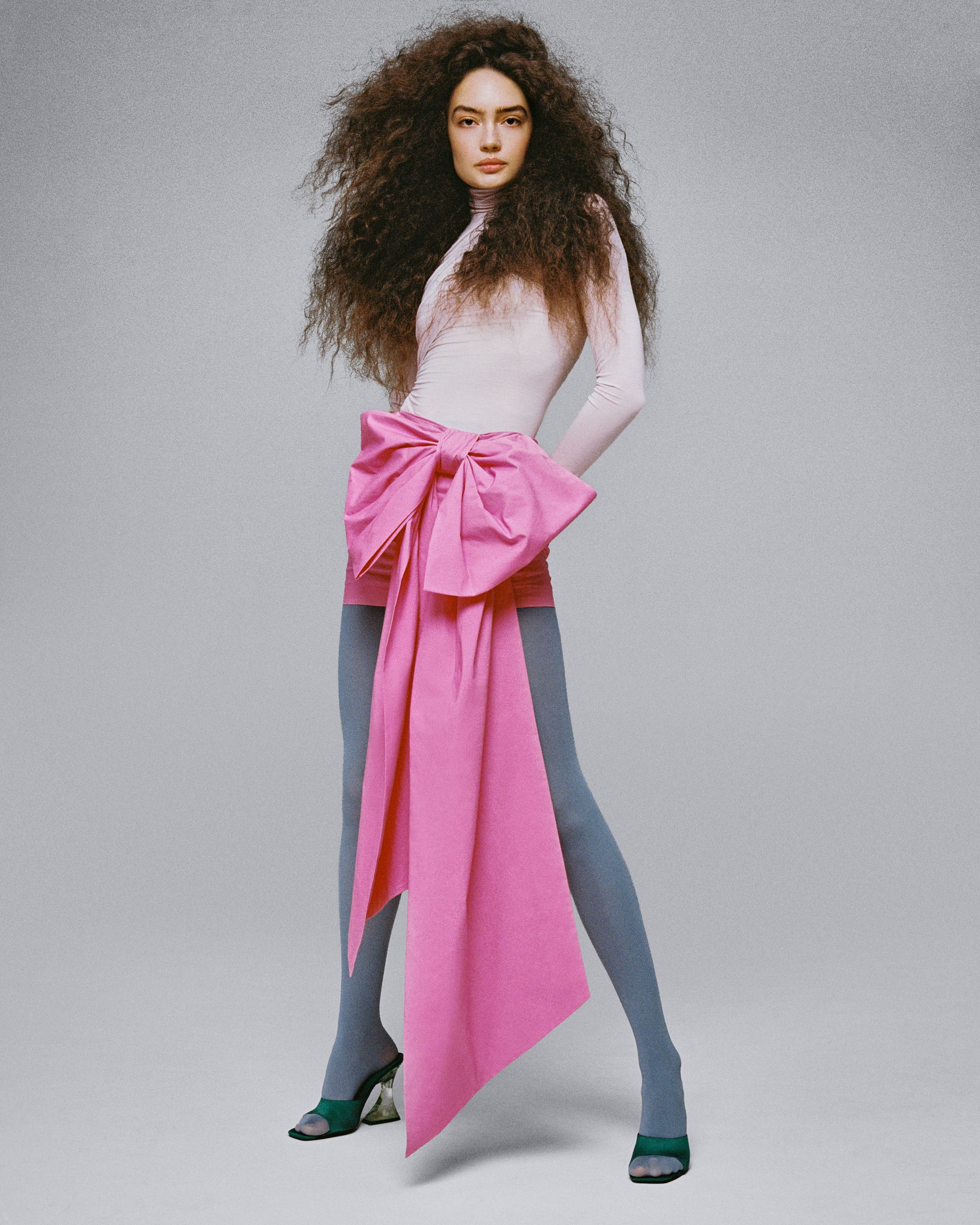 Sasha Kichigina in slate gray tights and a pale pink top with an oversized pink bow, standing against gray