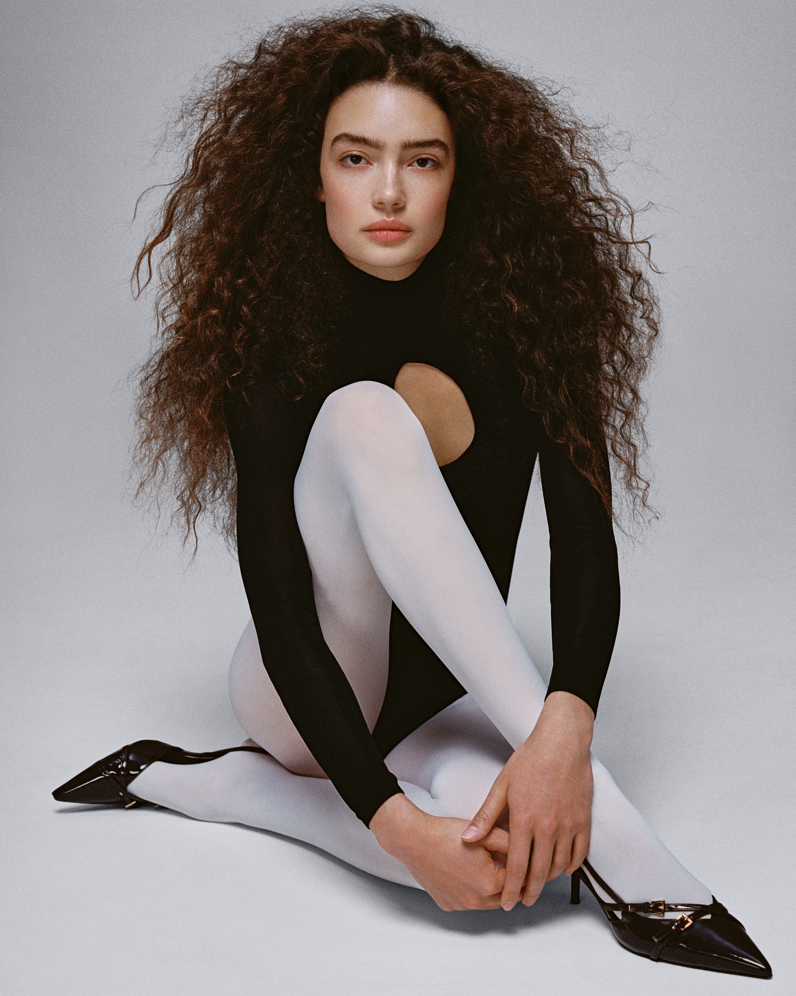 Sasha Kichigina hugging her knees in white tights, a black cutout top and black patent heels