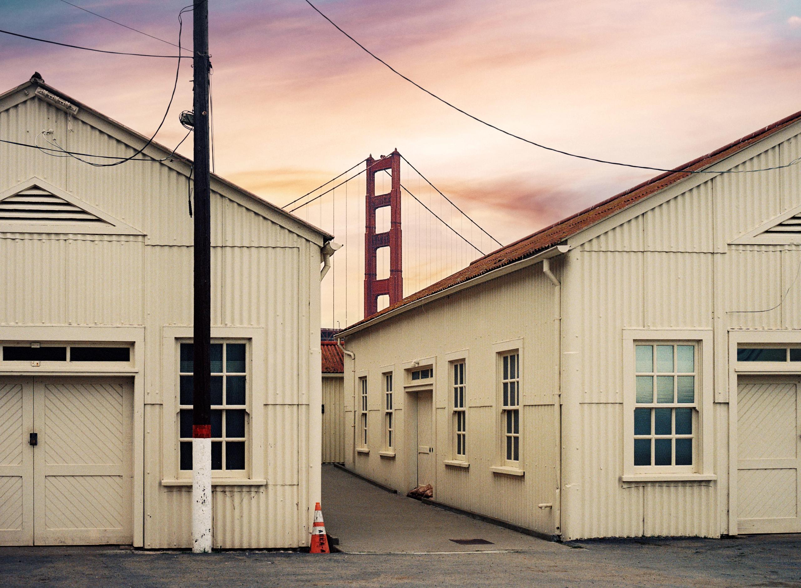 Pale wooden buildings in the Presidio at dusk, the Golden Gate Bridge tower rising behind