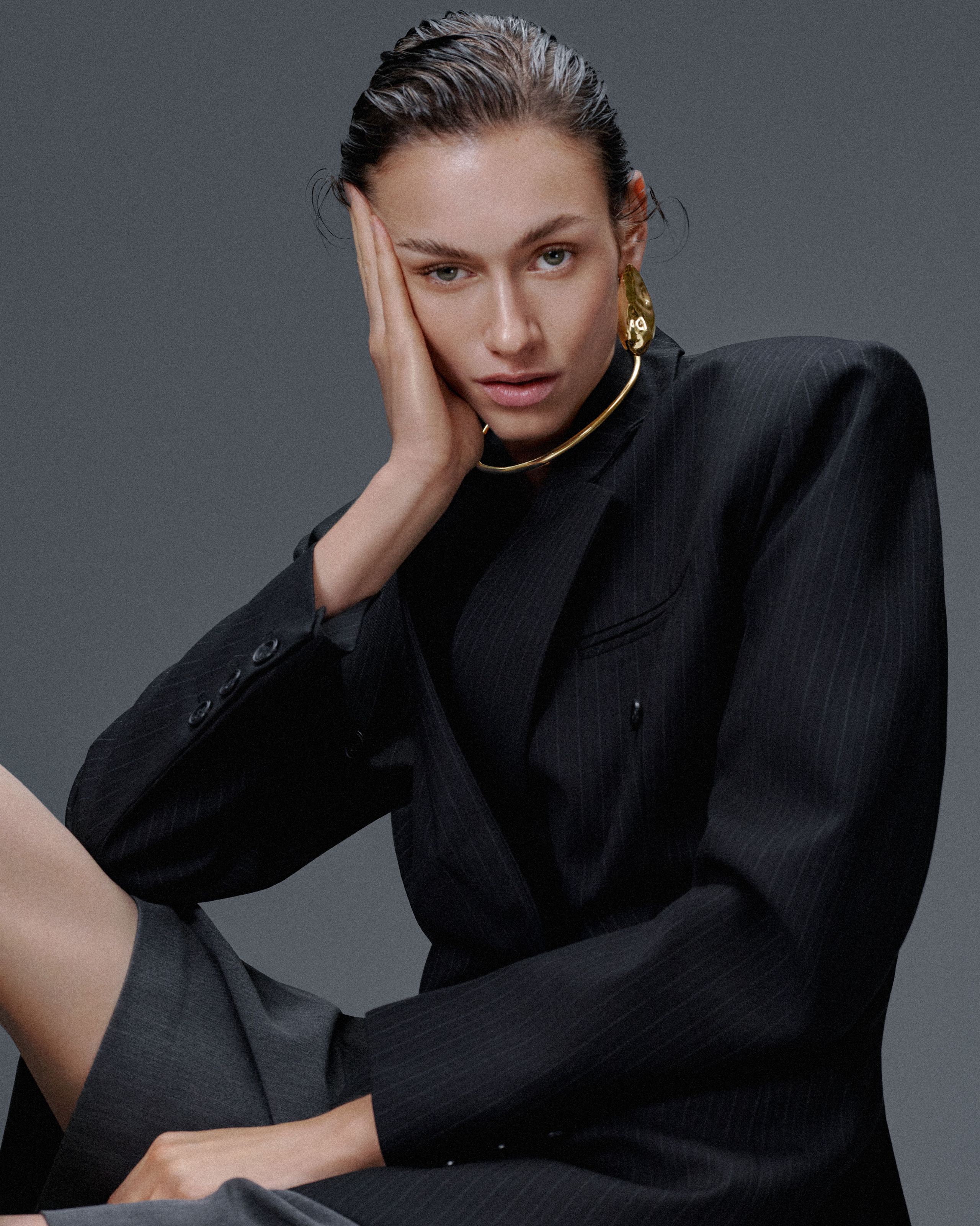 Model in a sculptural pinstripe blazer with slicked-back hair and bold Alexis Bittar jewelry, posing against a gray backdrop.