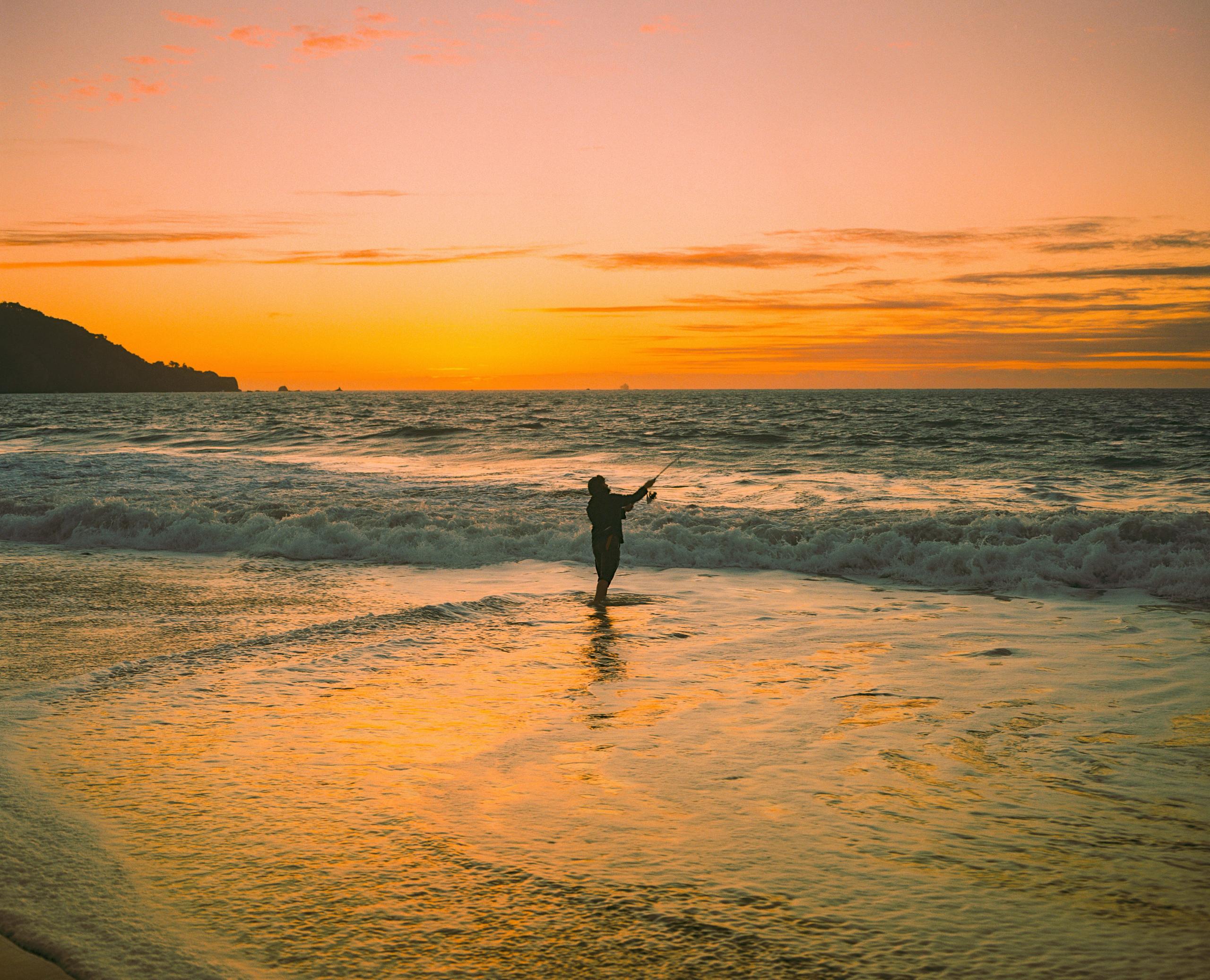 A silhouetted figure lifting an arm in the surf as the sky glows orange at sunset