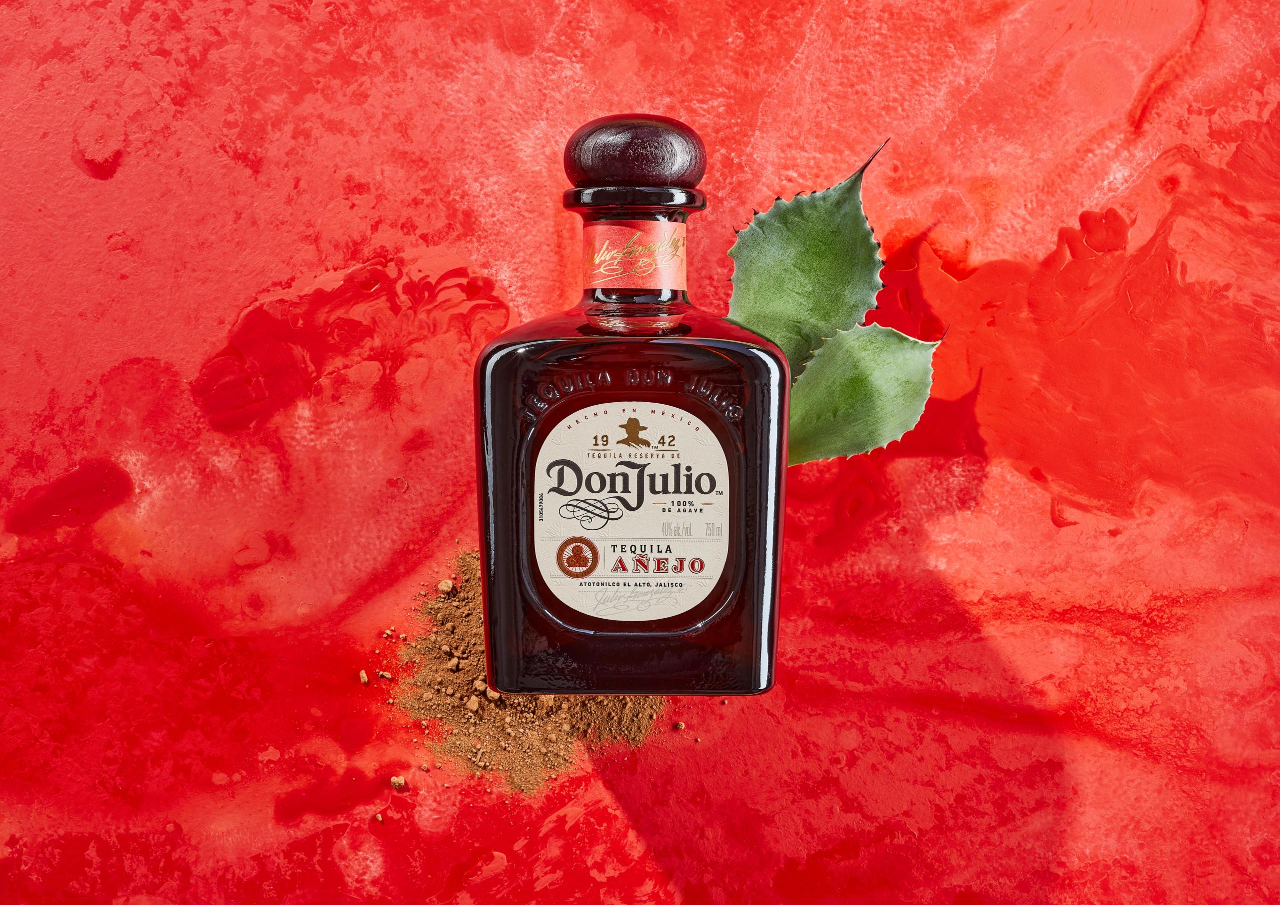 Overhead view of a Don Julio Añejo bottle on textured red, a sprig of agave and a dusting of ground spice beside it