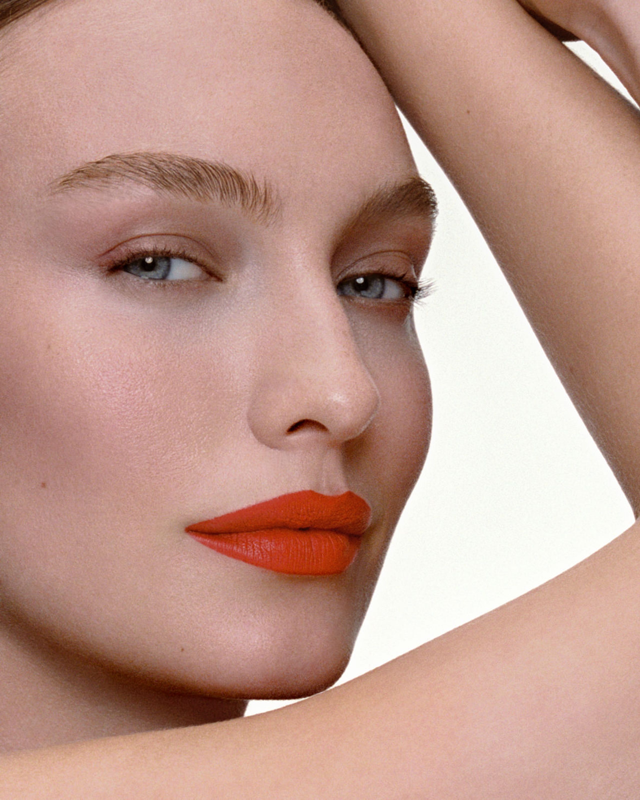 Model with an arm raised over her head, feathered brows and coral-orange lips