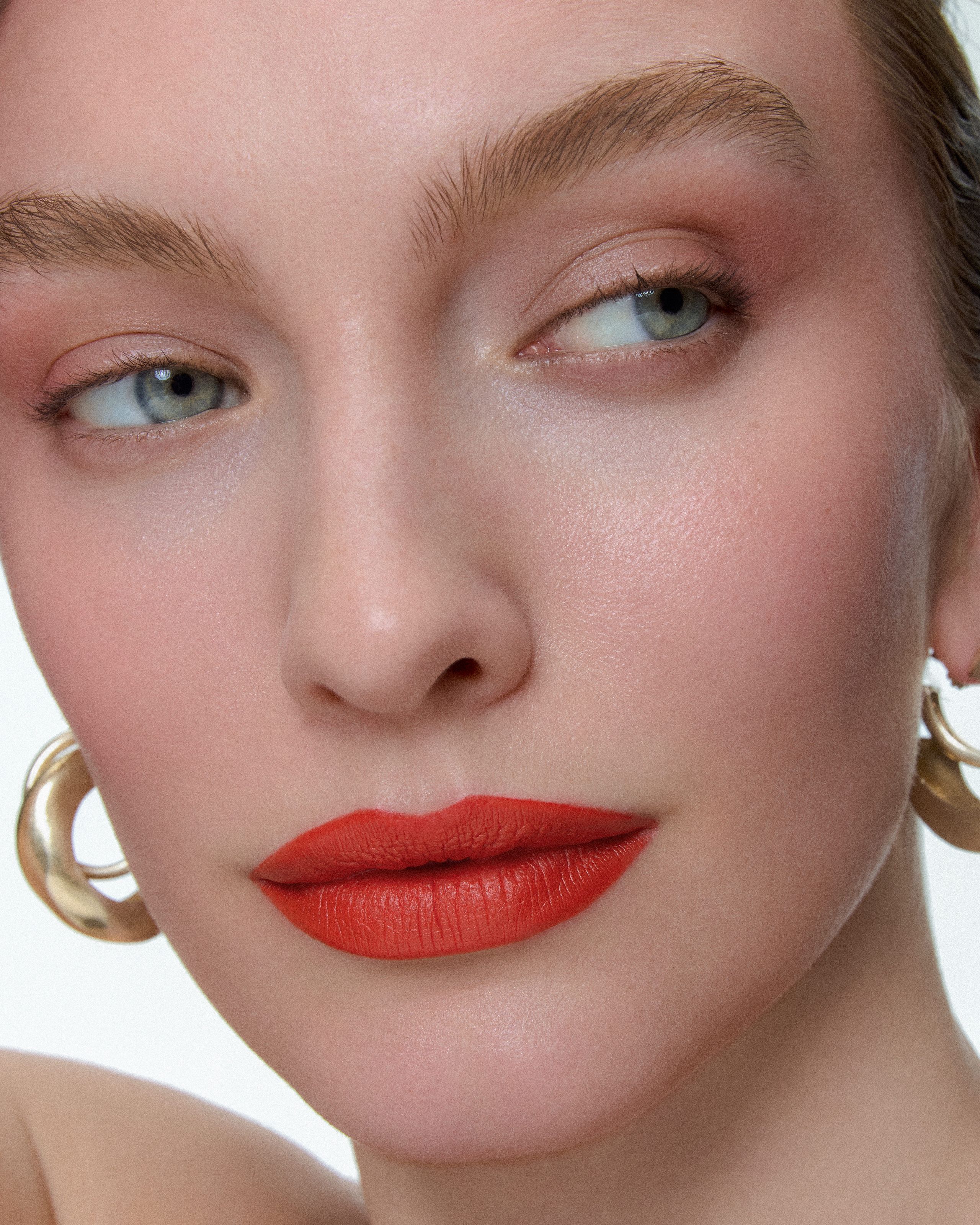 Model glancing sideways with orange-red lips and sculptural gold AGMES hoop earrings