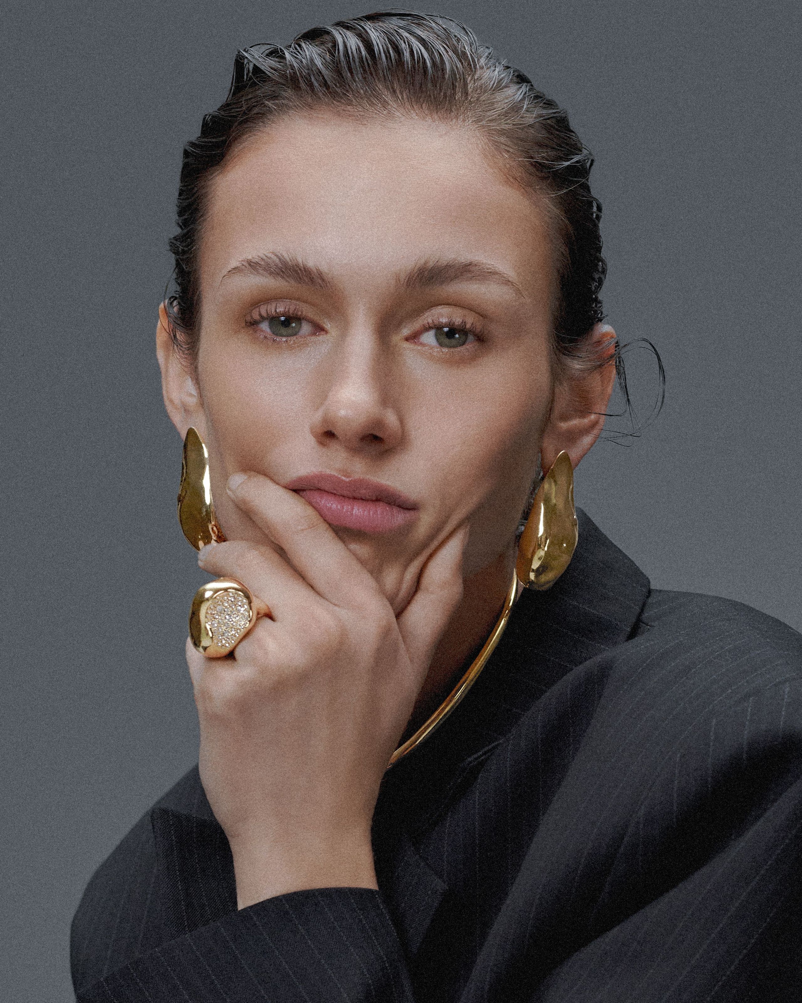 Model in a pinstripe blazer wearing sculptural gold Alexis Bittar earrings and ring, posed against a gray backdrop.