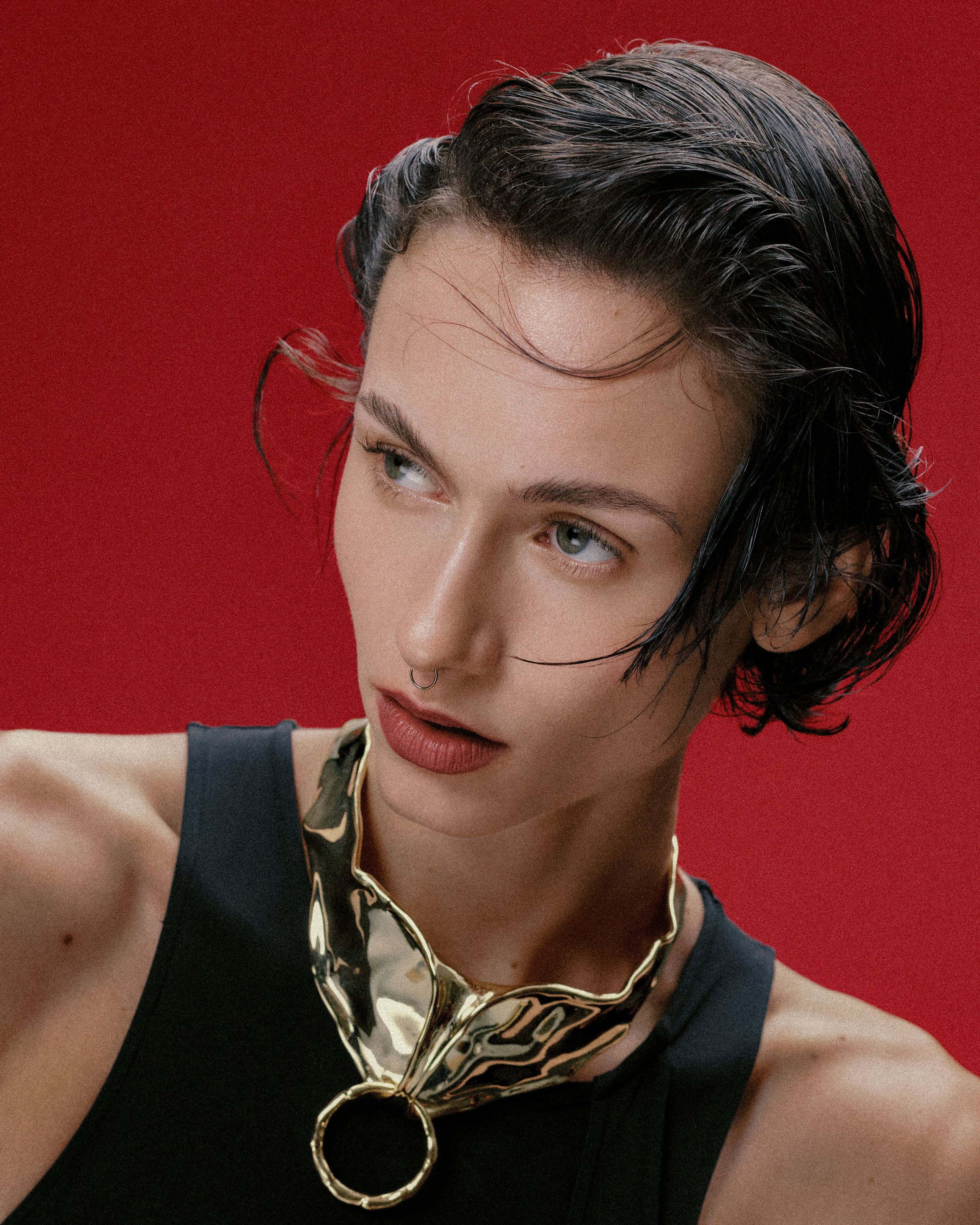 Model wears a sculptural gold statement necklace with wet-look hair against a vivid red backdrop.