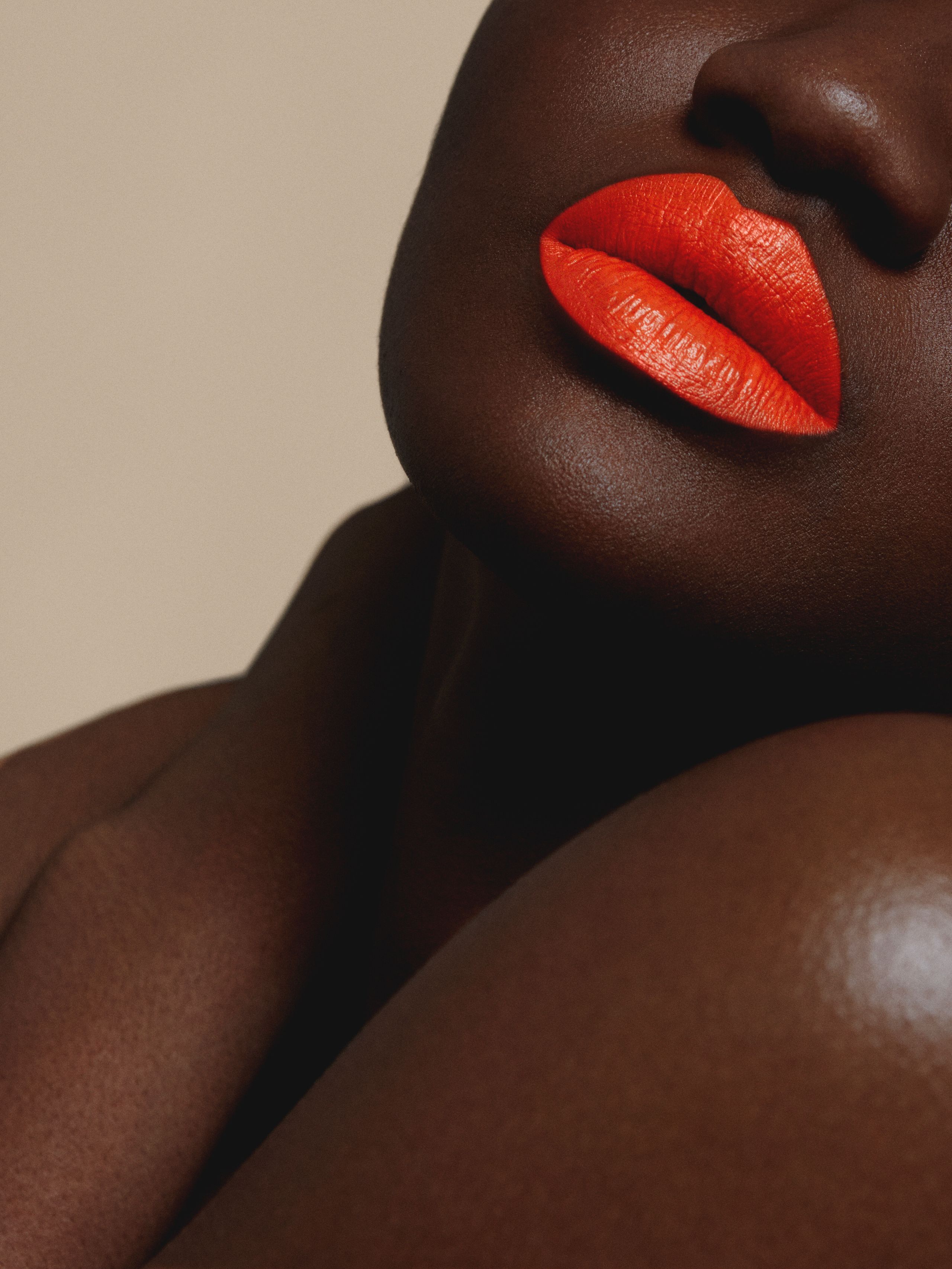 Ayet Betty resting her chin on a bare shoulder, orange lips catching soft light against a beige backdrop