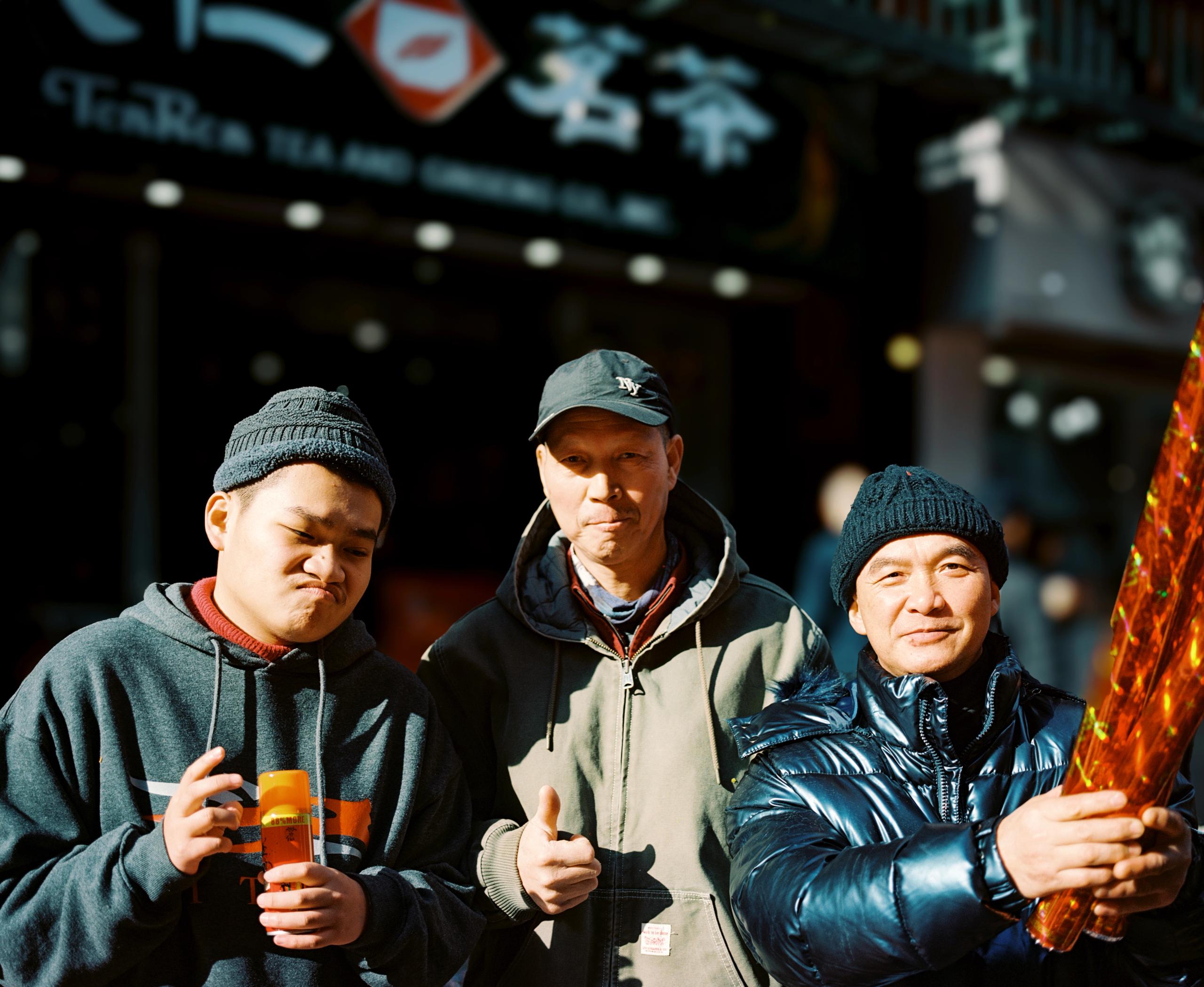 Three men in winter caps smiling outside a shop, one holding a string of firecrackers