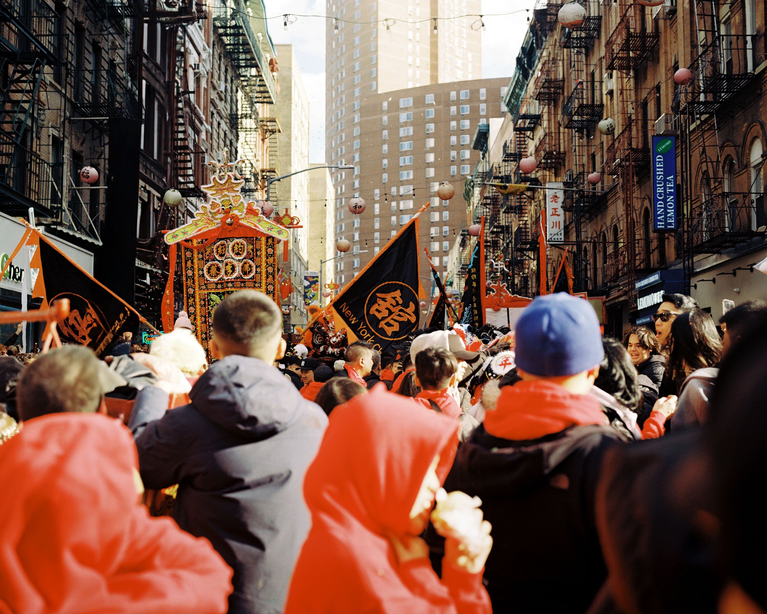 Parade banners and lion dancers above a crowd on a narrow Chinatown street lined with fire escapes