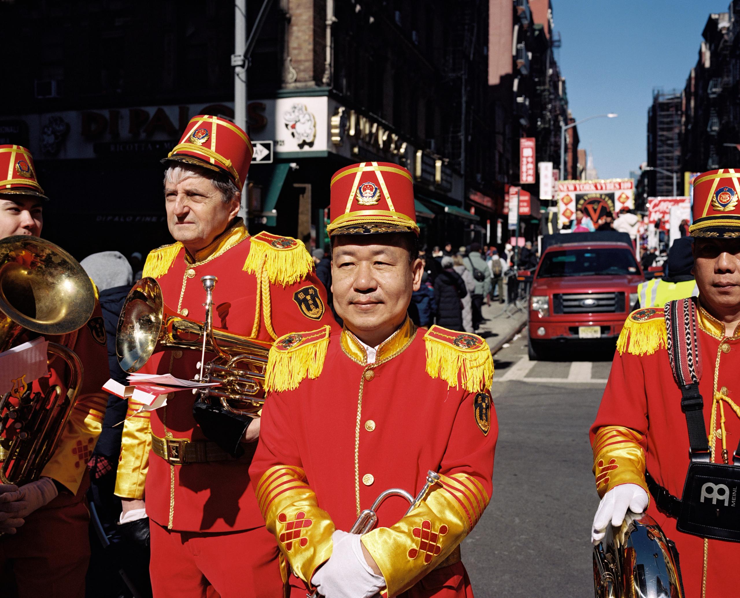 Marching band members in red and gold uniforms and plumed caps on a Chinatown street during a Lunar New Year parade
