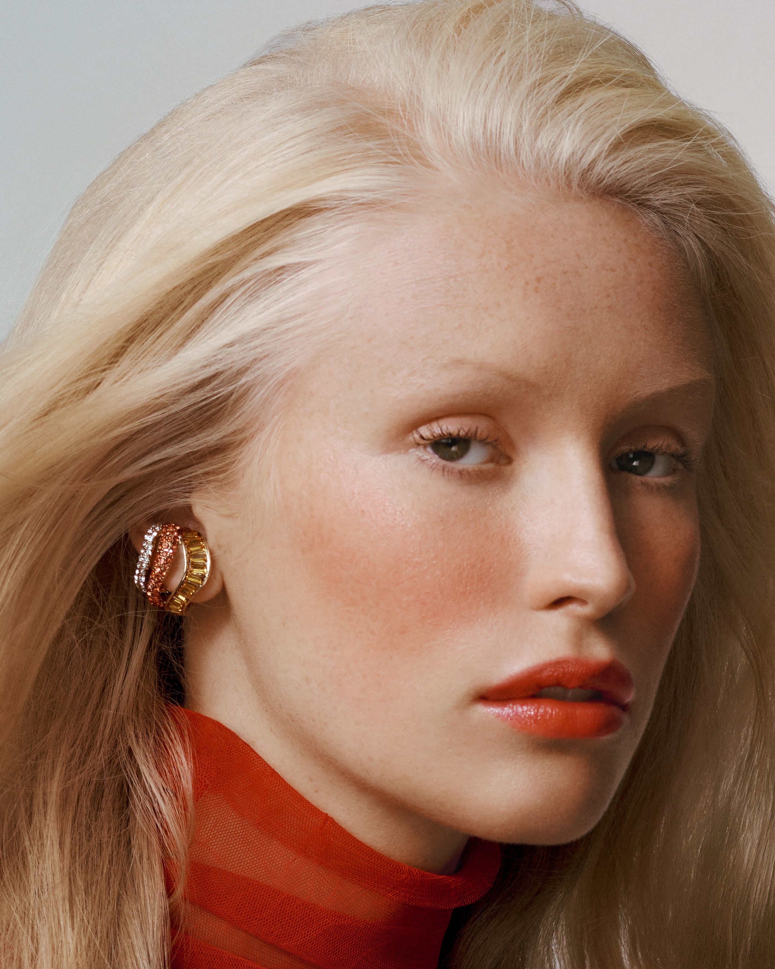 Close-up of a blonde model with coral lips, a sculptural gold earring and a sheer red turtleneck
