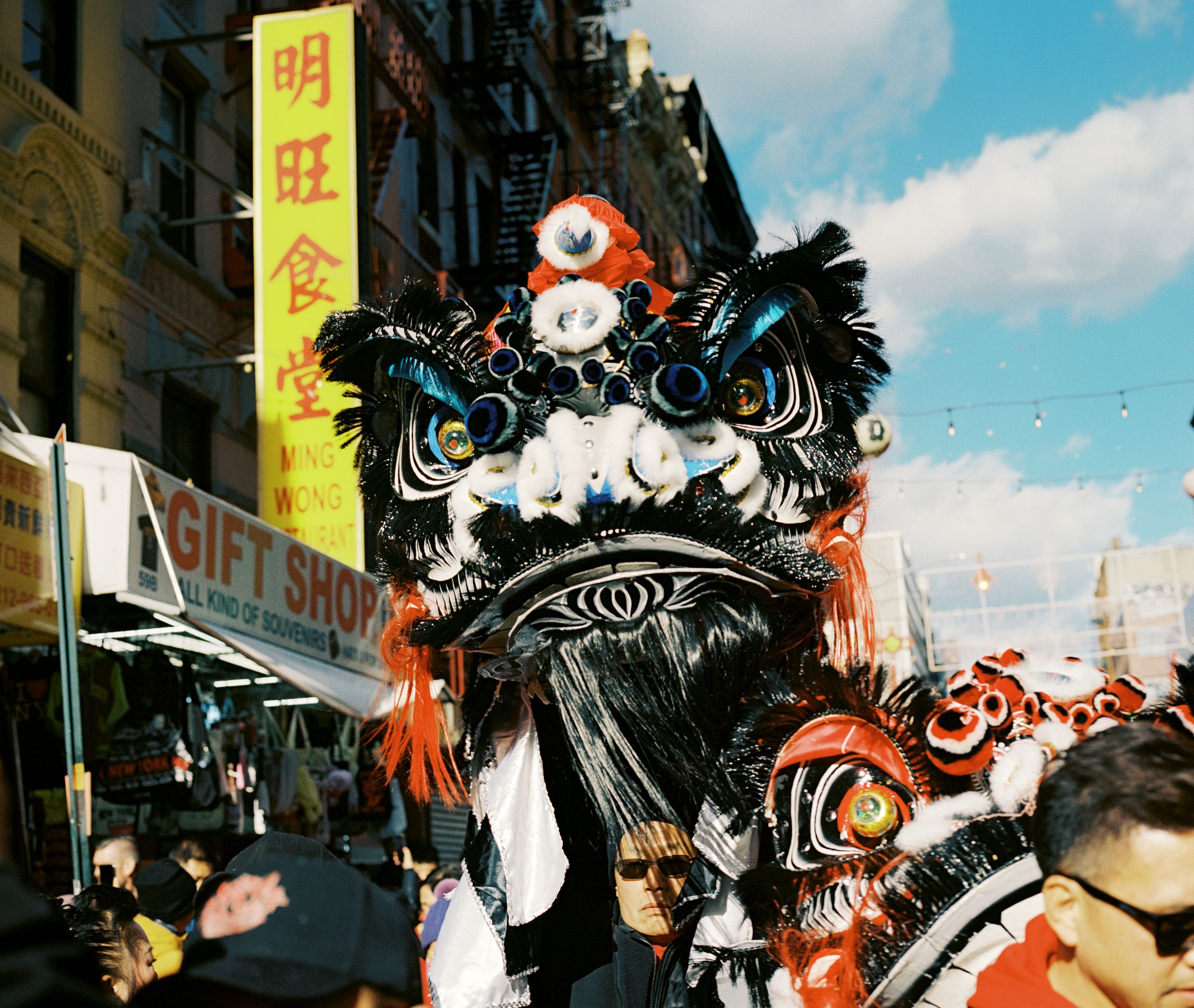 Black and white lion dance head raised over a crowd beneath a yellow restaurant sign