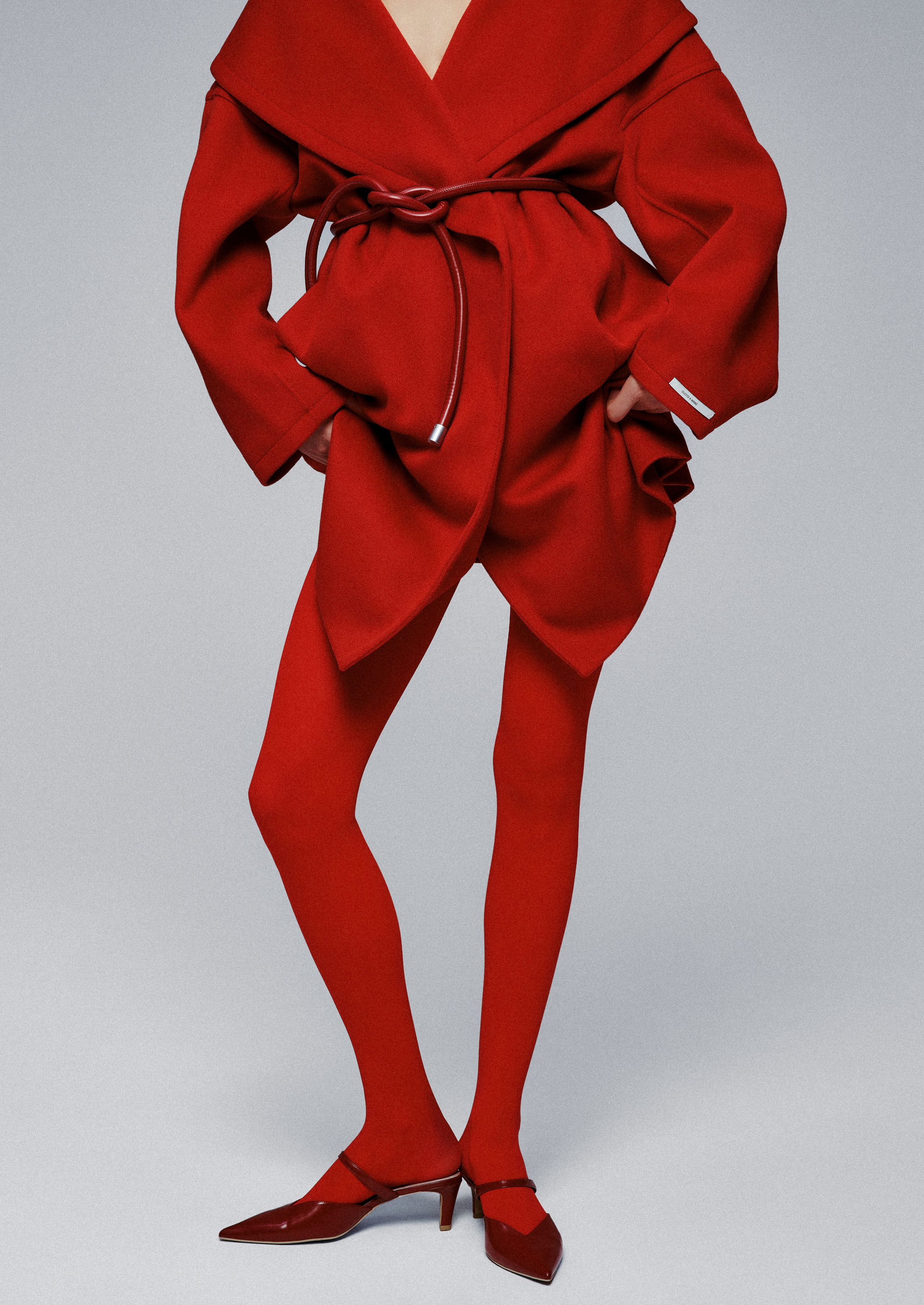 Sasha Kichigina in red tights, a belted red wrap coat and red kitten heels, cropped at the shoulders against gray