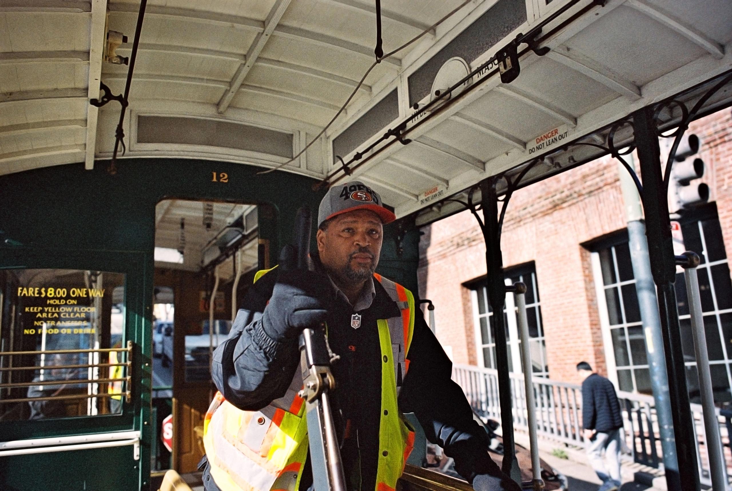 A cable car gripman in a safety vest and cap at the controls, looking into the camera