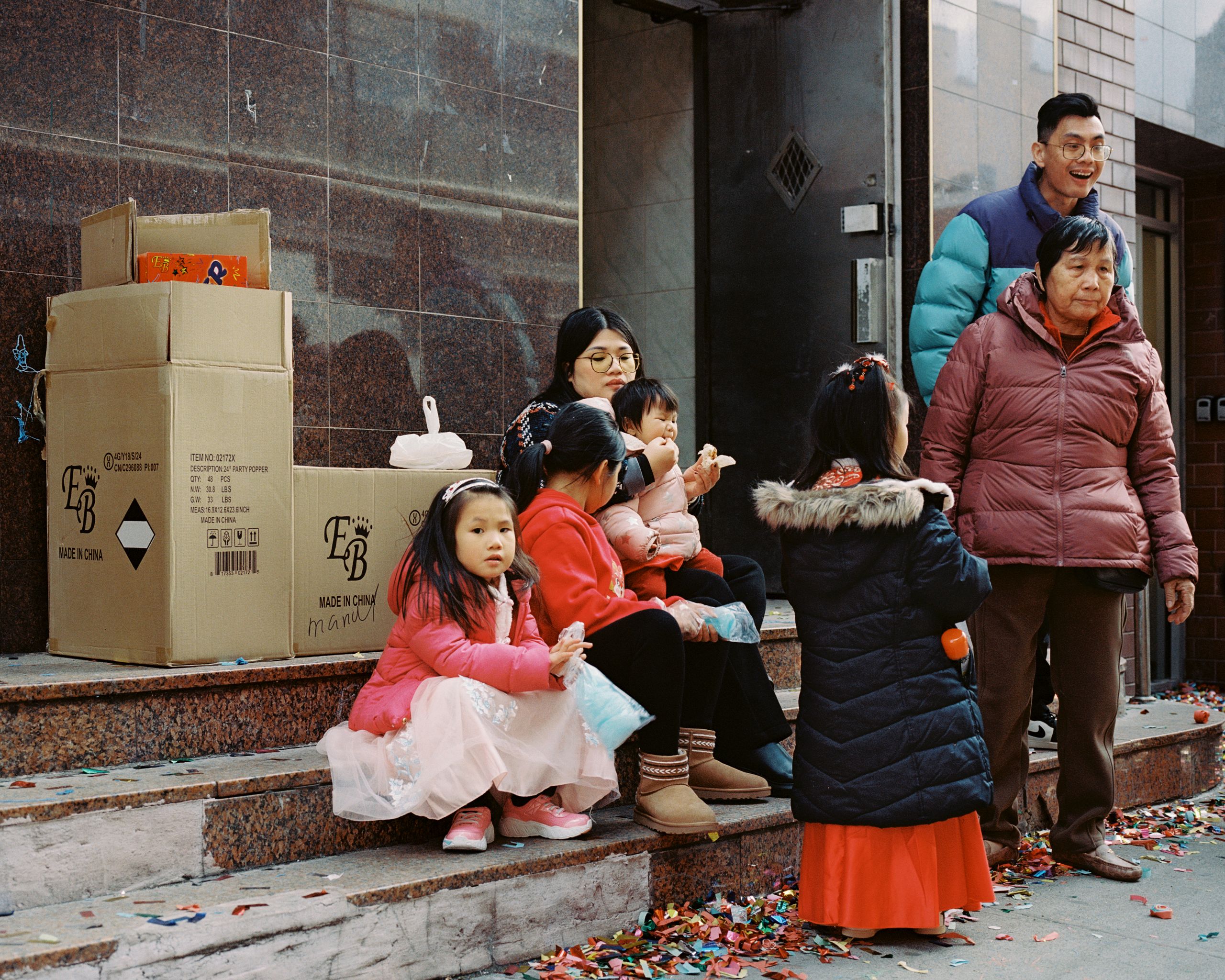 Families resting on stoop steps beside stacked boxes as the Lunar New Year crowd passes