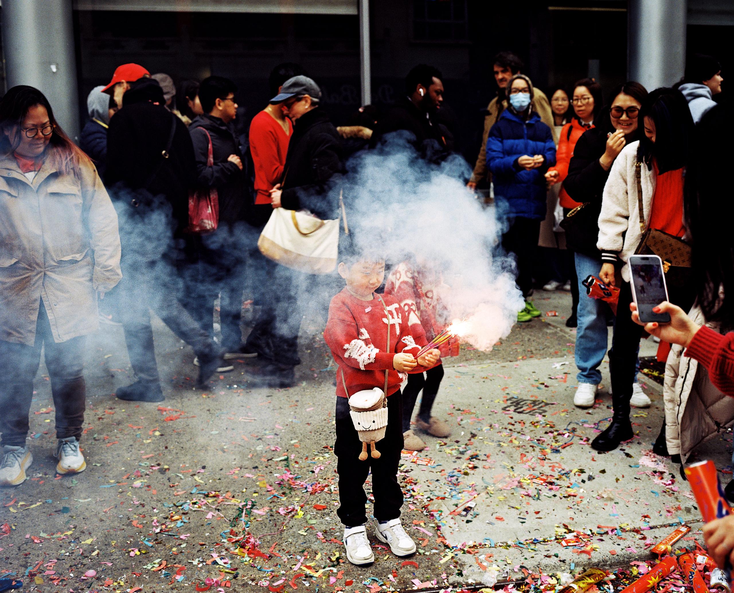 A child in a red jacket holds a smoking firecracker as a Lunar New Year crowd looks on, red paper scattered underfoot