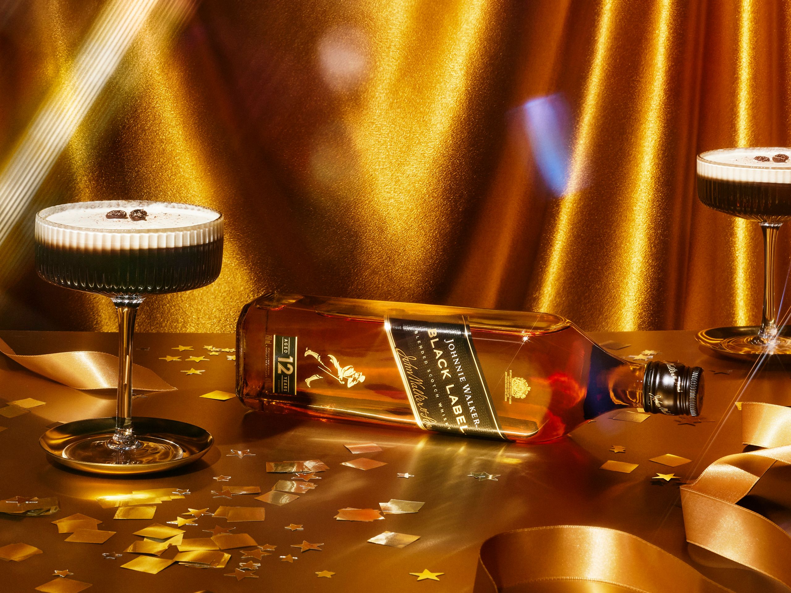 Johnnie Walker Black Label laid on its side between two foam-topped cocktails against draped gold satin
