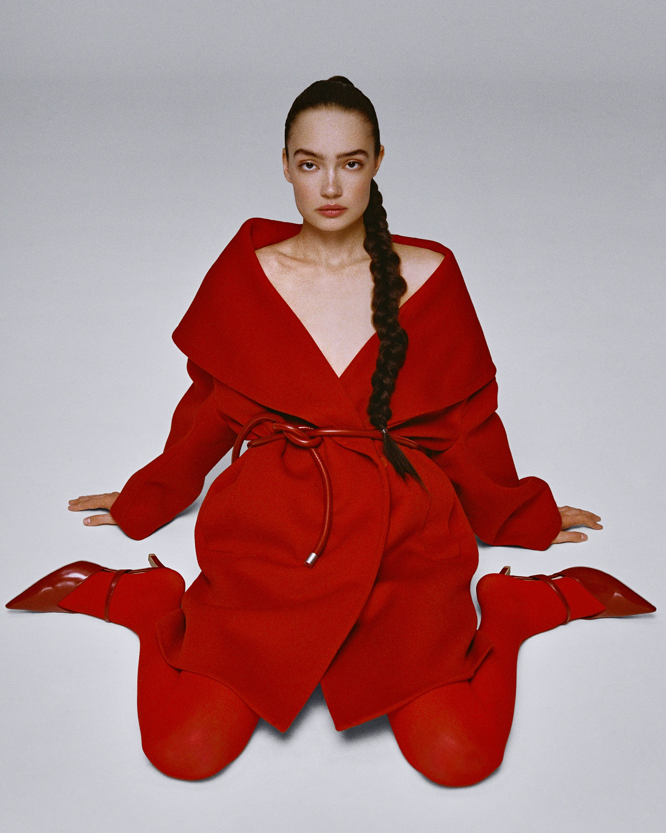 Sasha Kichigina kneeling in red tights and an oversized red wrap coat, a long braid over her shoulder