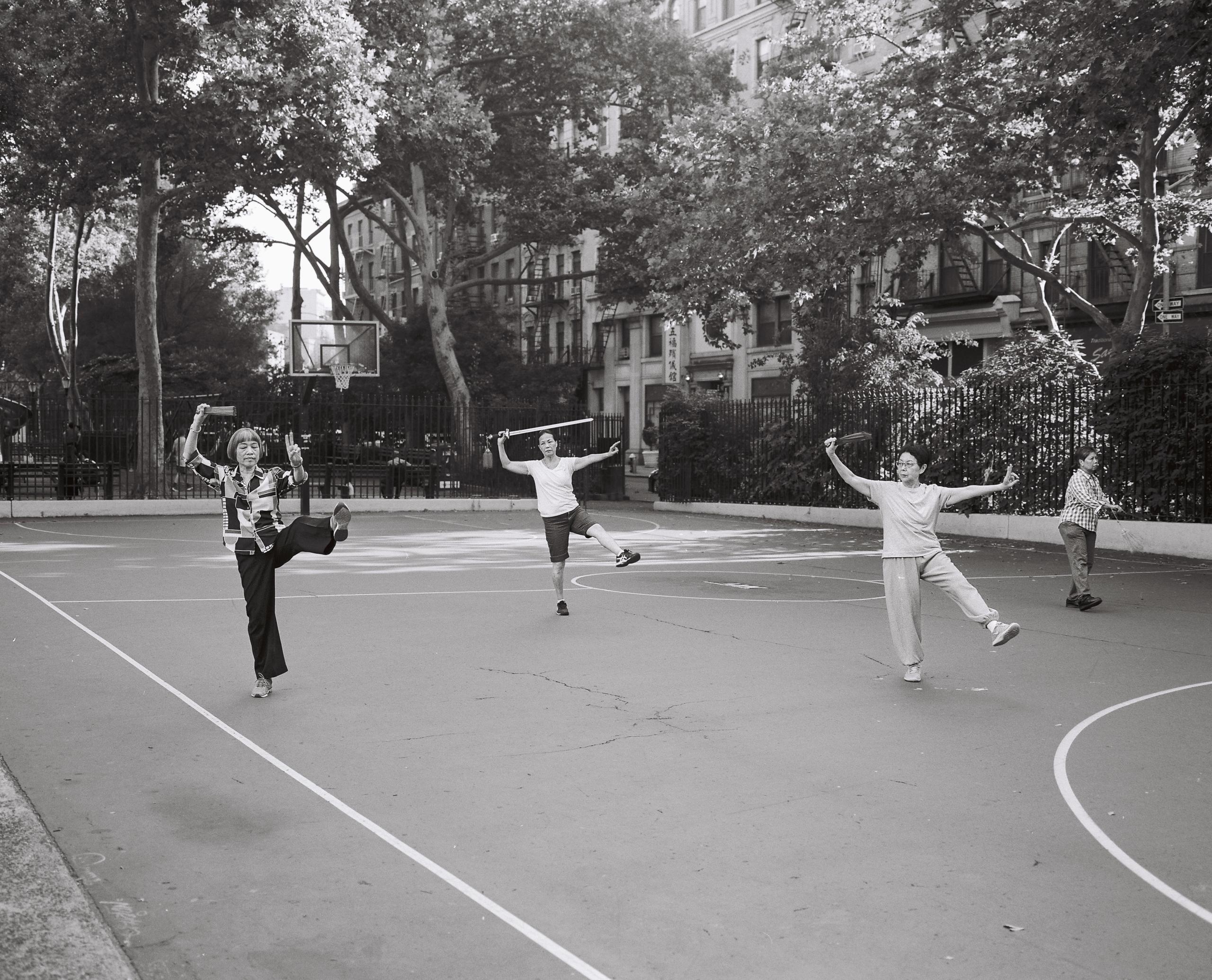 Older women practicing morning exercises on a park basketball court beneath tall trees, black-and-white film