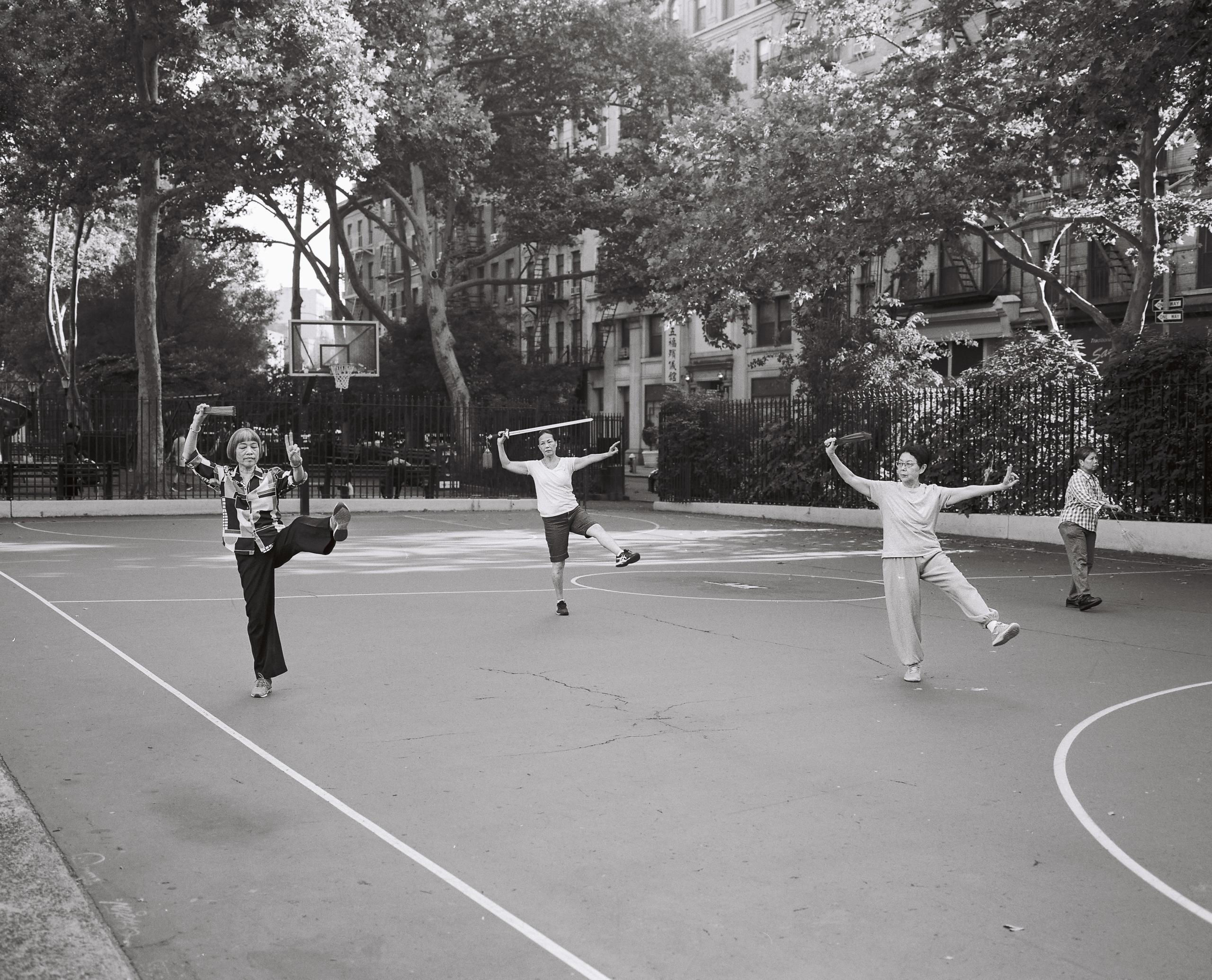 Older women practicing morning exercises on a park basketball court beneath tall trees, black-and-white film