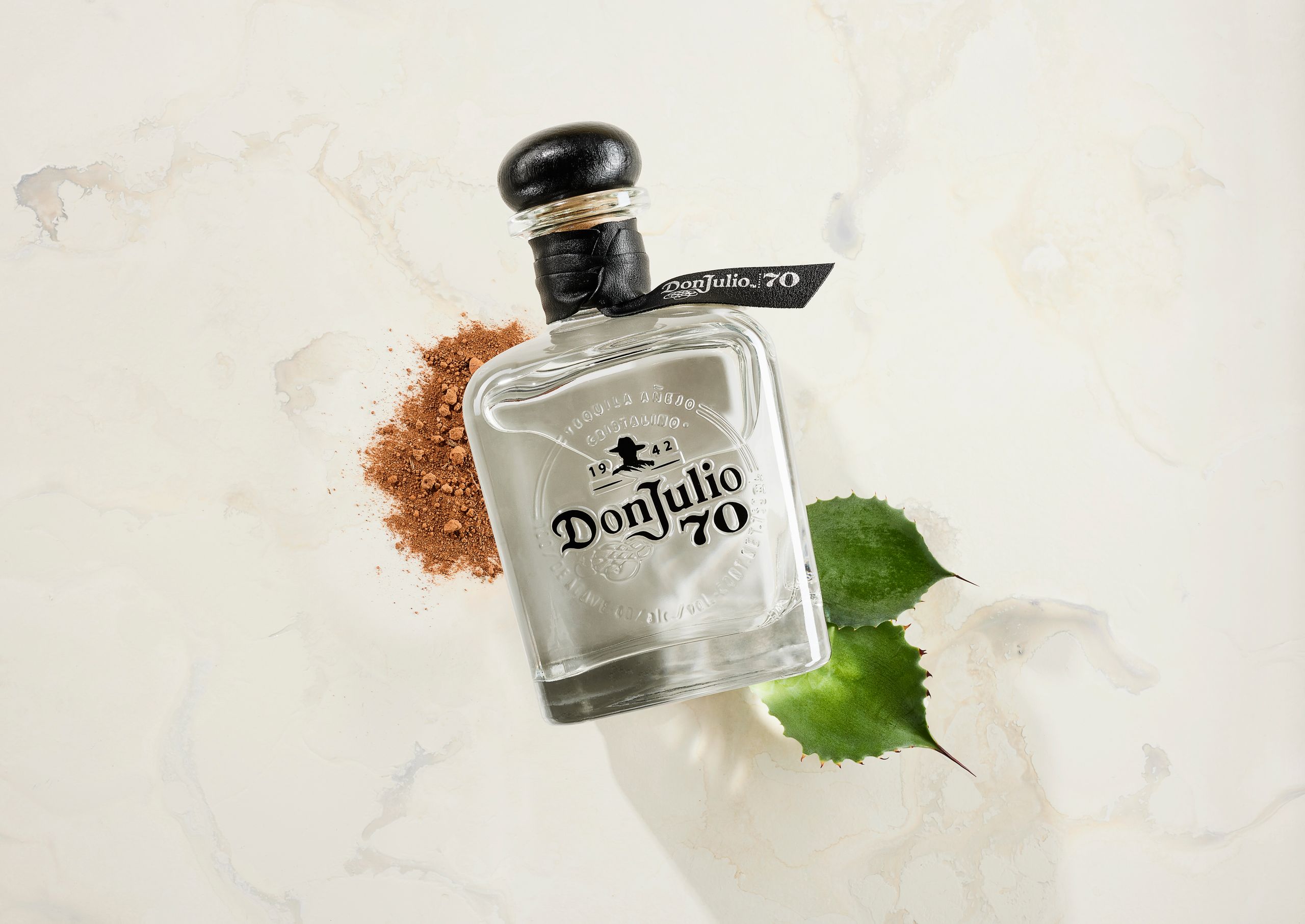 Clear Don Julio 70 Añejo bottle on a pale backdrop with green leaves and cocoa powder