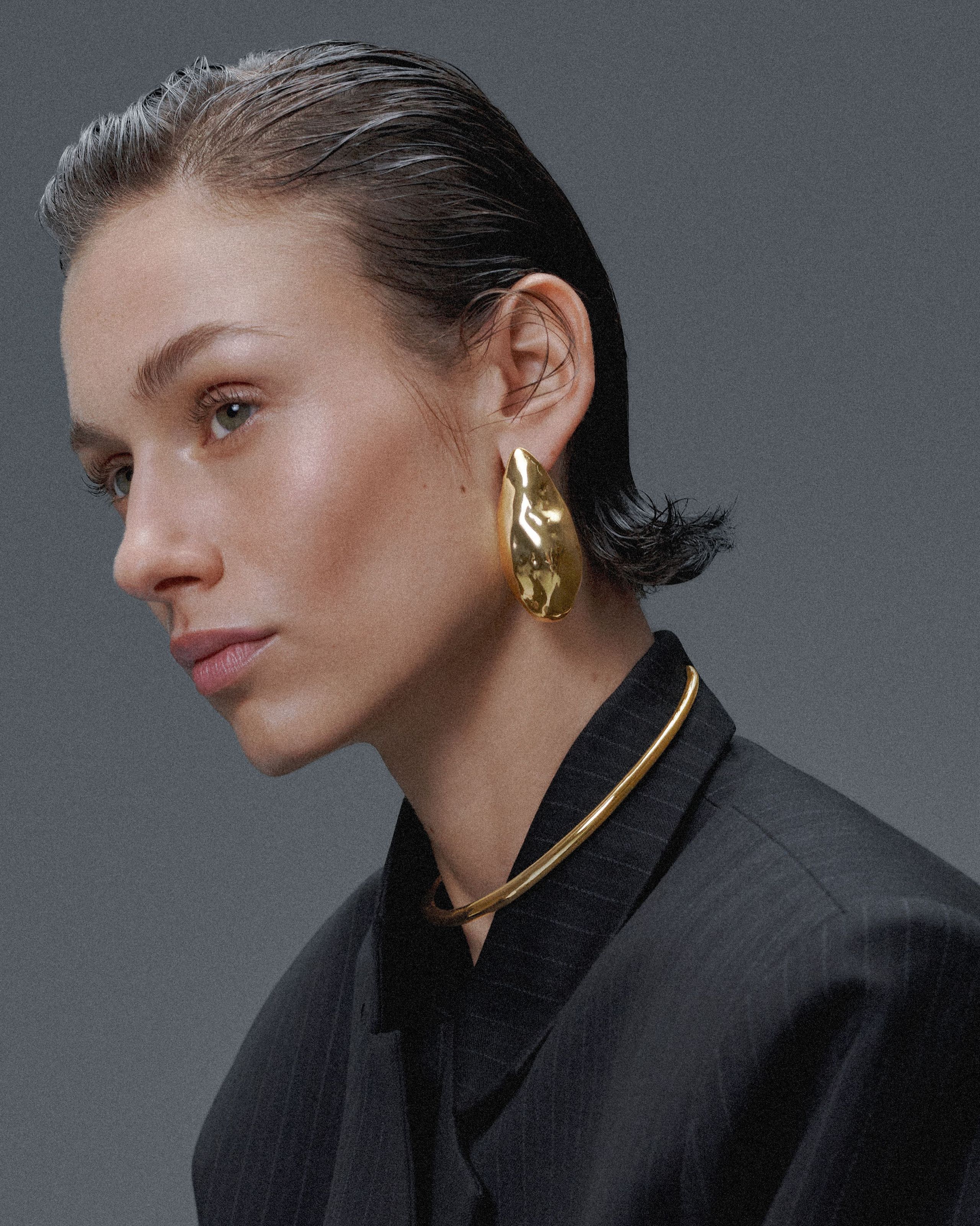 Model in a pinstripe blazer with slicked-back hair, wearing sculptural gold Alexis Bittar earrings and necklace.