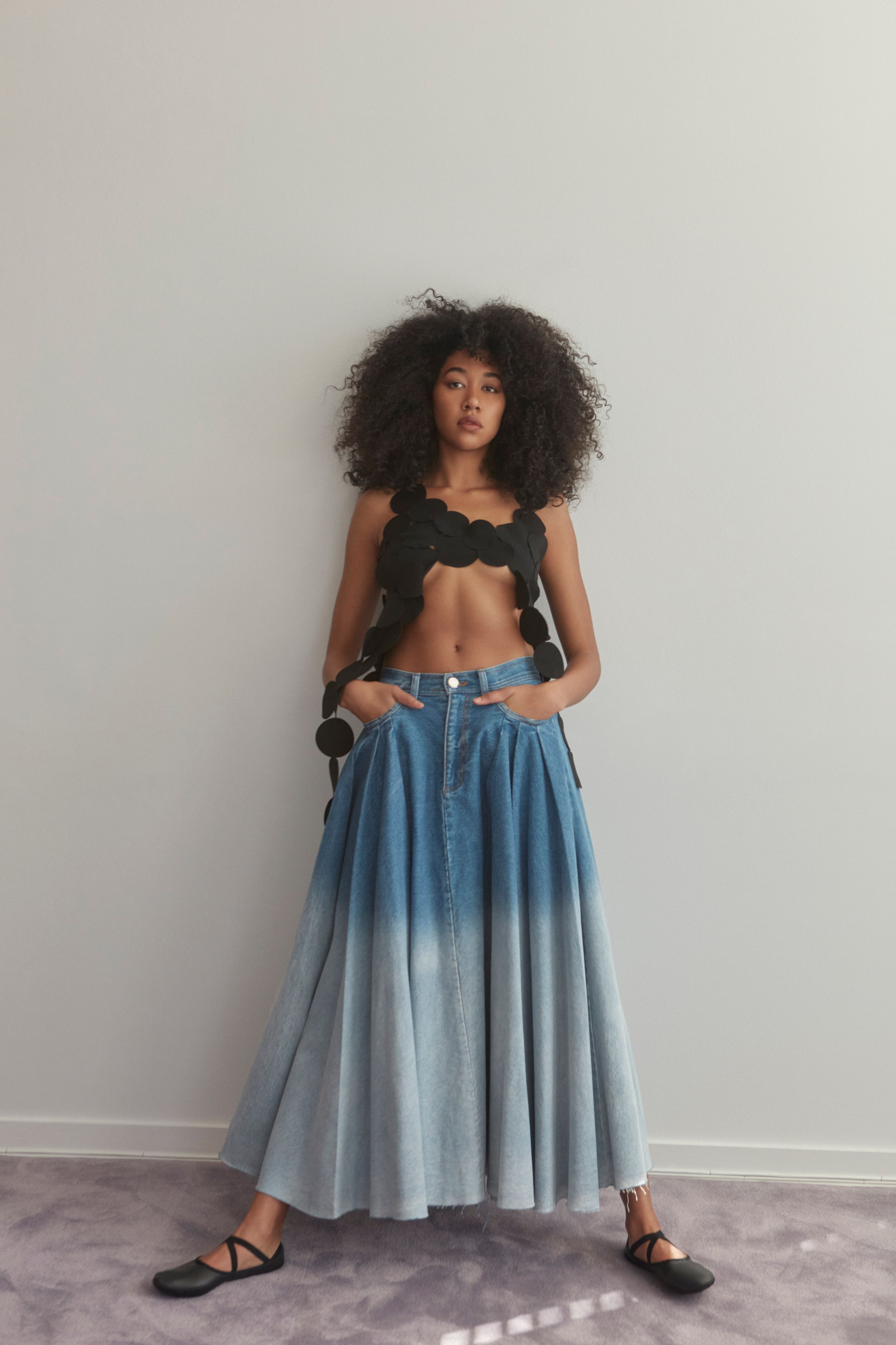 Aoki Lee Simmons against a pale wall in a sculptural black top and long ombré denim skirt, hands in pockets