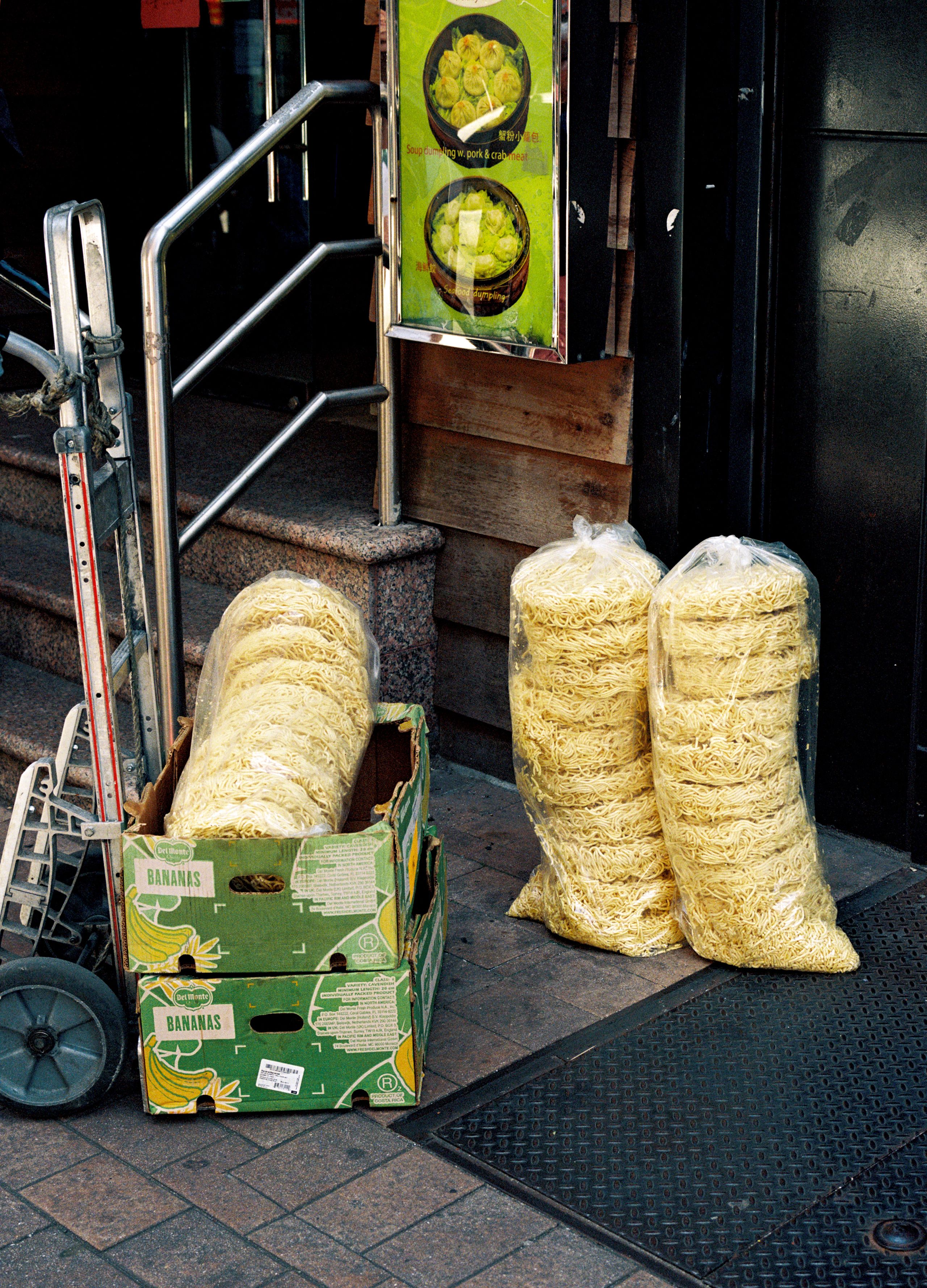 Bundles of dried noodles stacked on cardboard boxes outside a Chinatown storefront, a hand truck beside them