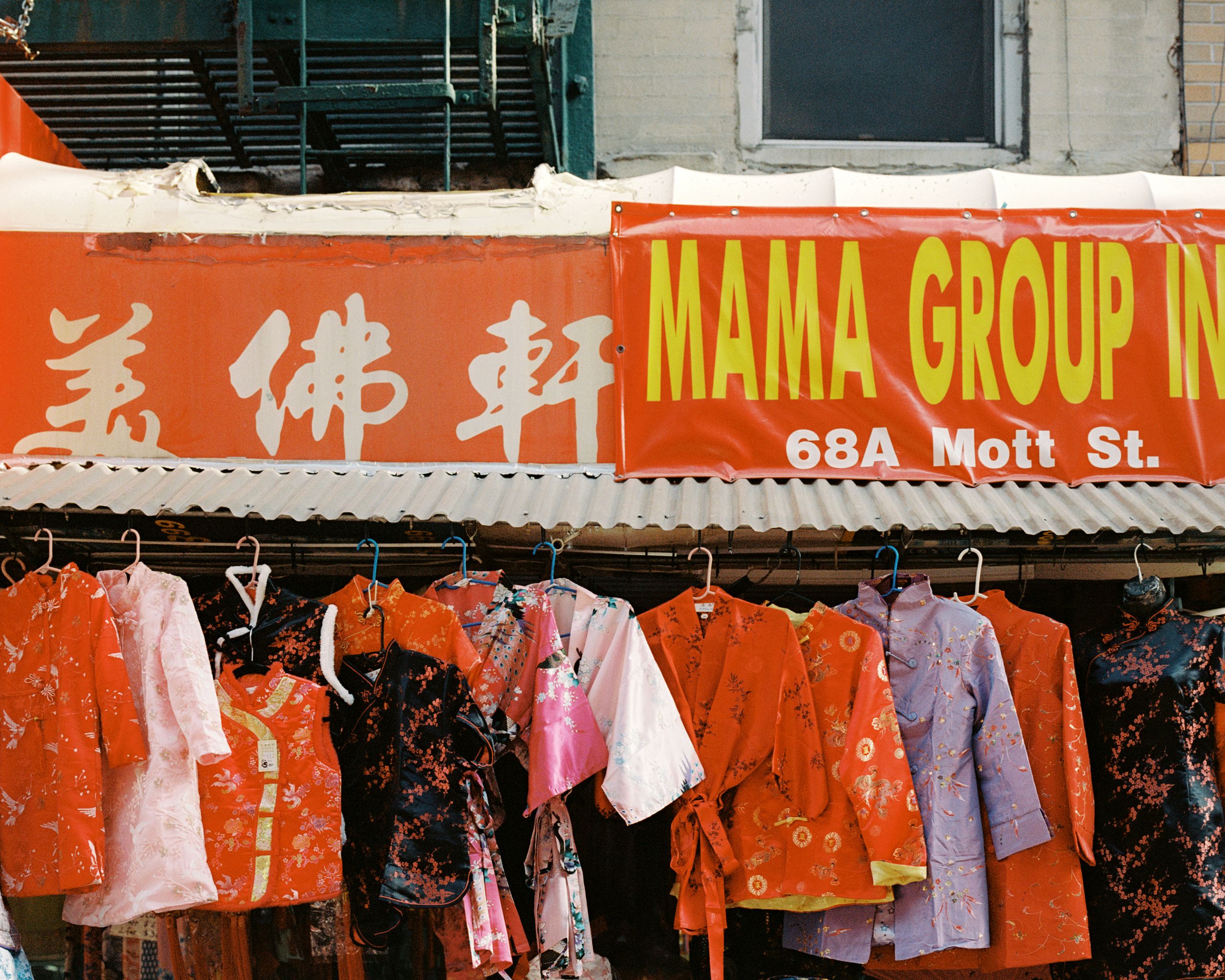 Red qipao and children’s outfits hanging beneath a shop sign on Mott Street