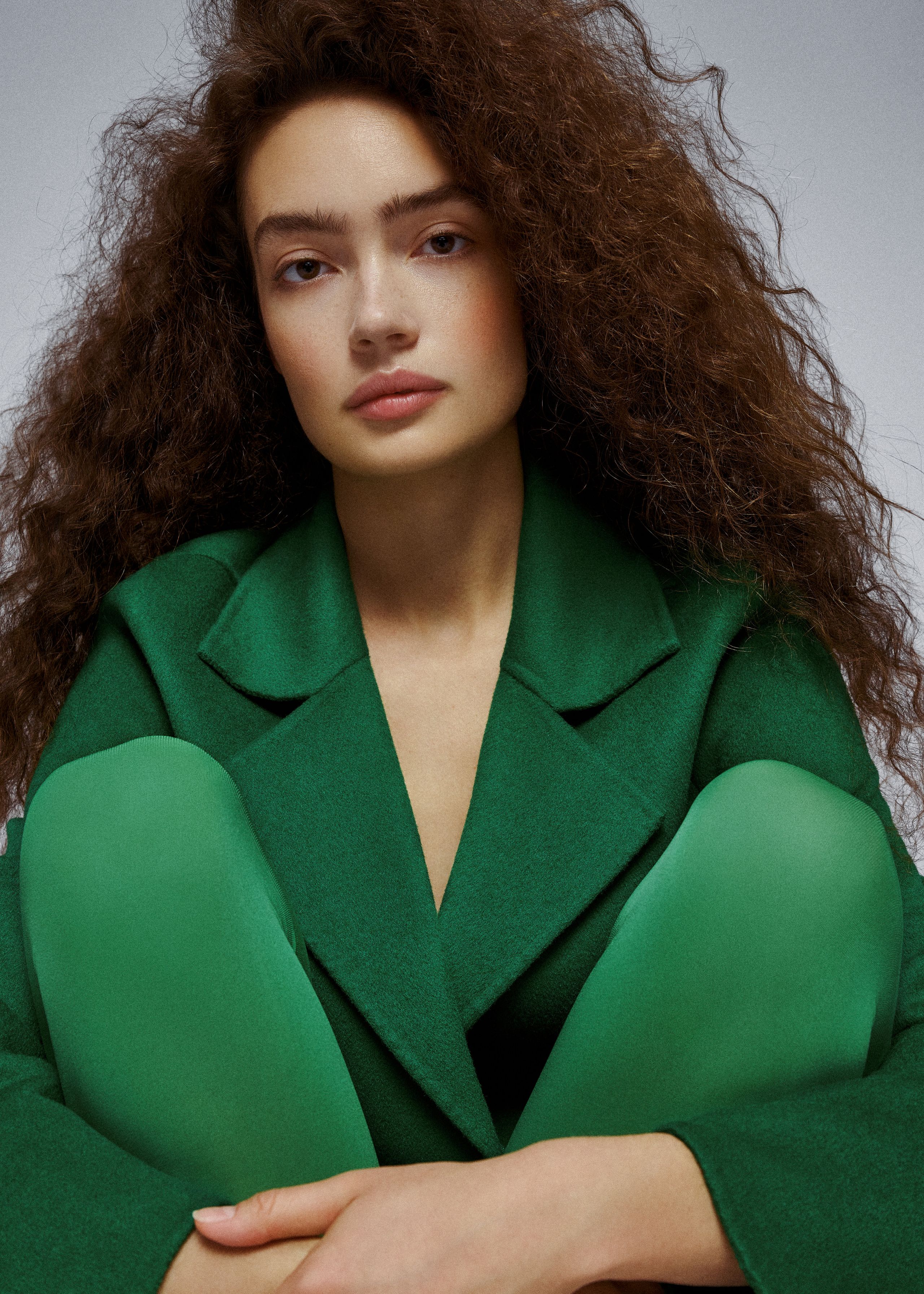 Close-up of Sasha Kichigina in a green coat, knees drawn up in matching green tights, soft studio light