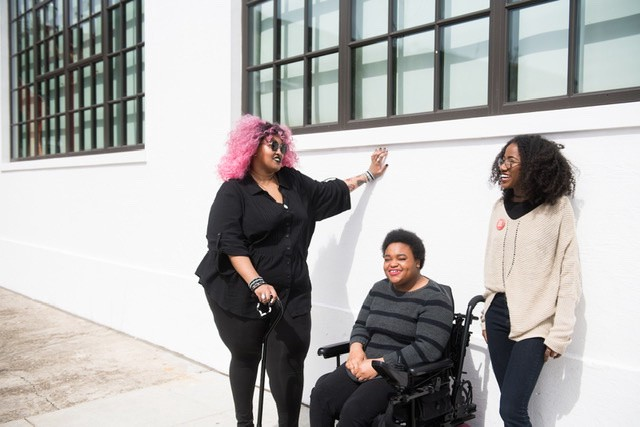 A group of people smile and chat up against a wall