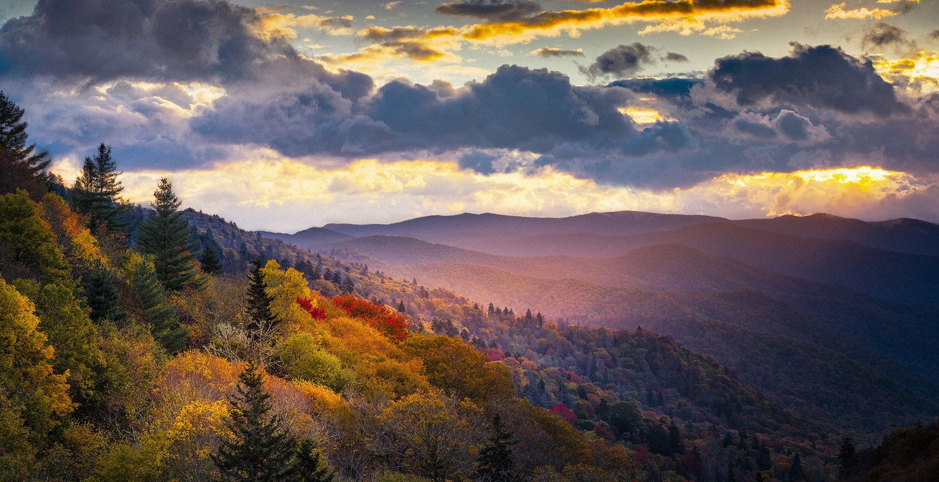 Sun beginning to set on a skyline of mountains and trees.