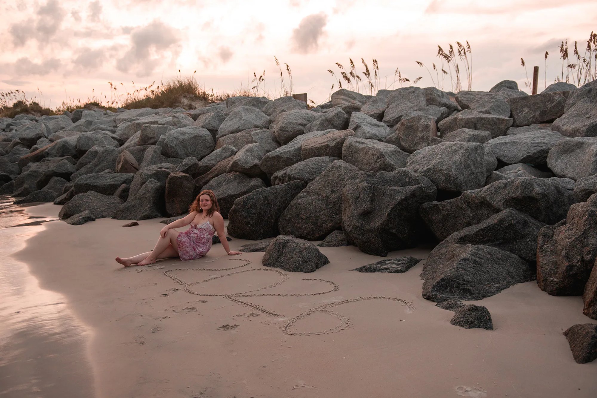 Senior photo session at Ponce Inlet, Nelly sitting on the sand near large rocks with “2025” written in the sand at sunset.