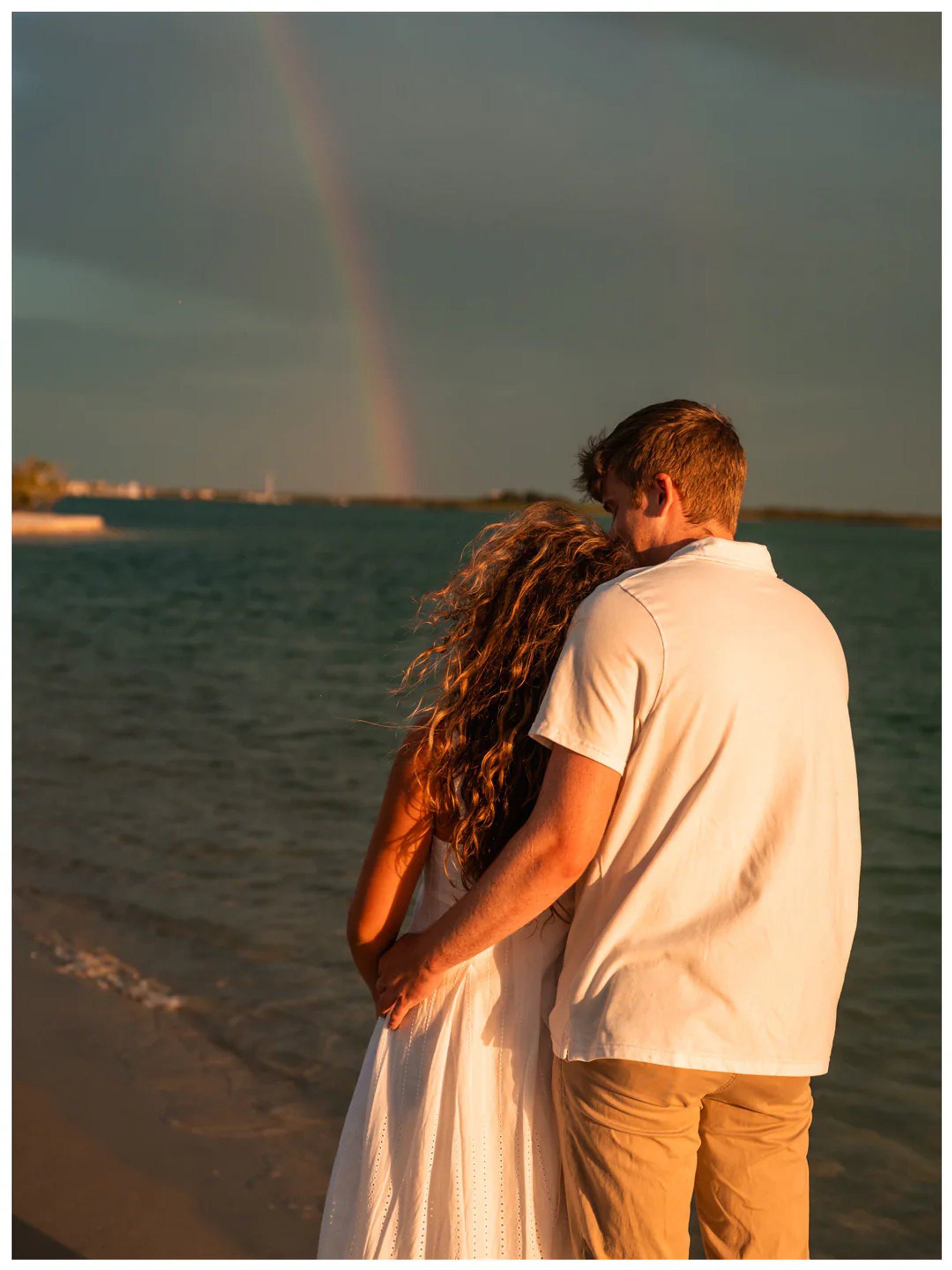 Couple embracing beside the water with a rainbow in the distance.