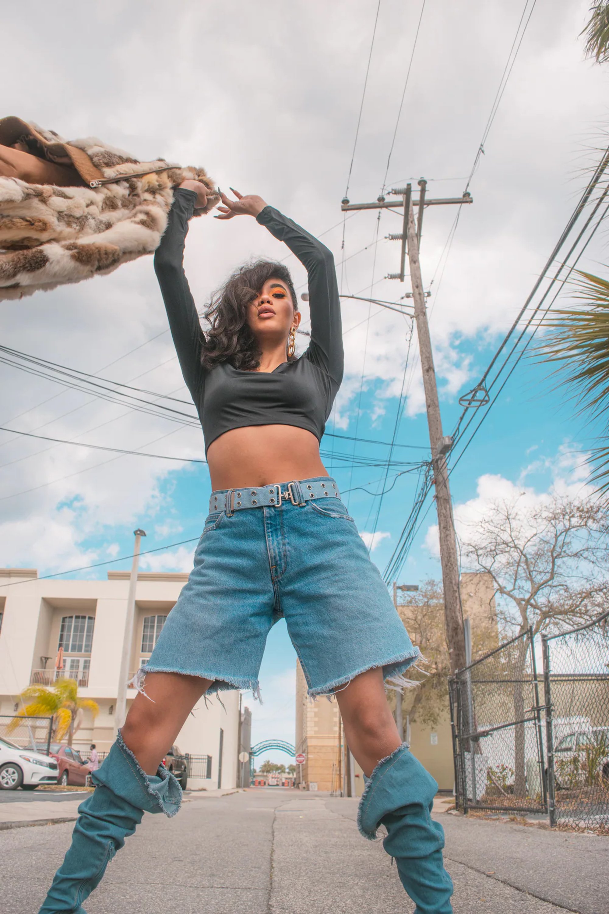 Woman raising her arms during a street-style portrait in denim shorts and blue boots.
