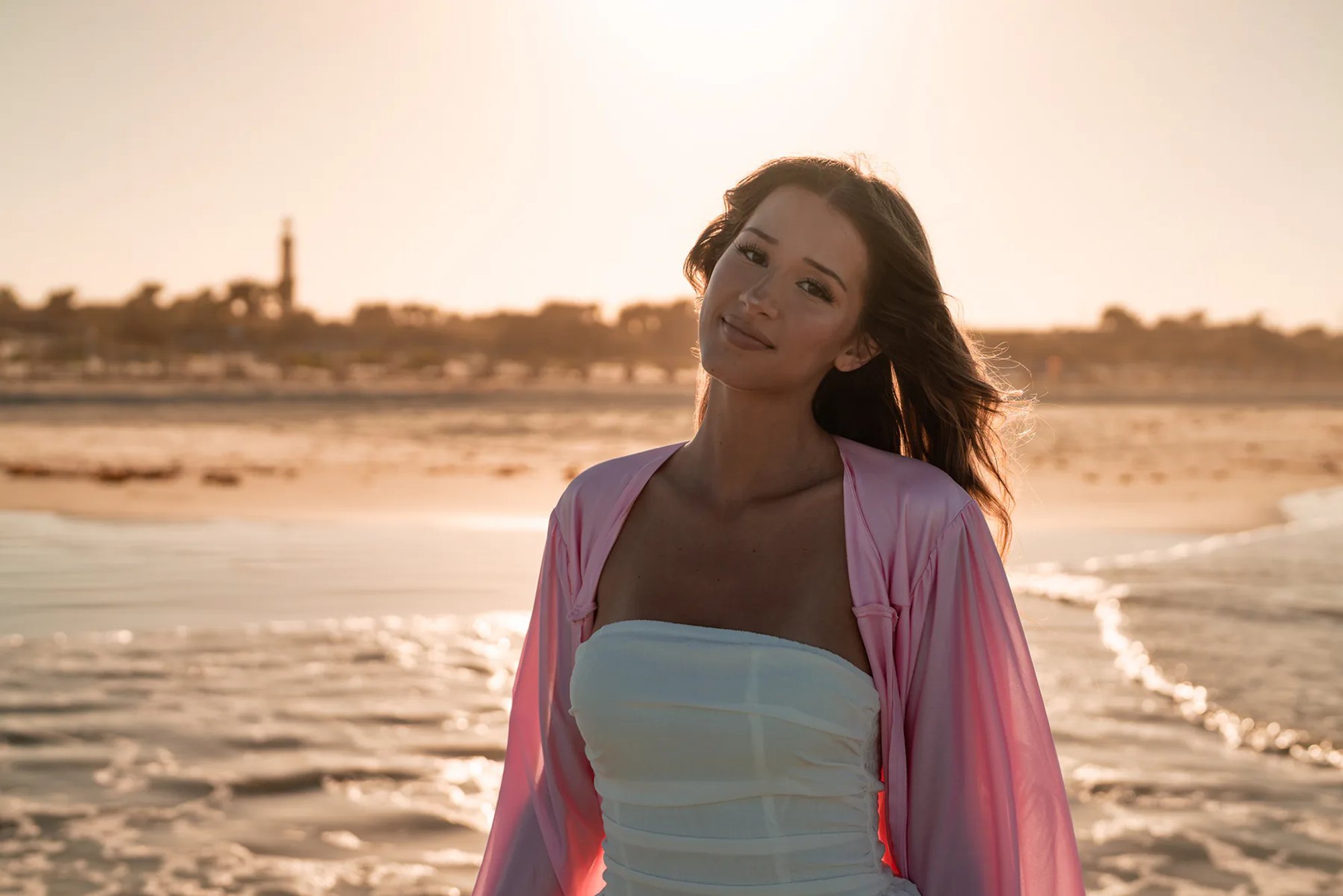 Young woman in a pink wrap standing beside the ocean at sunset.